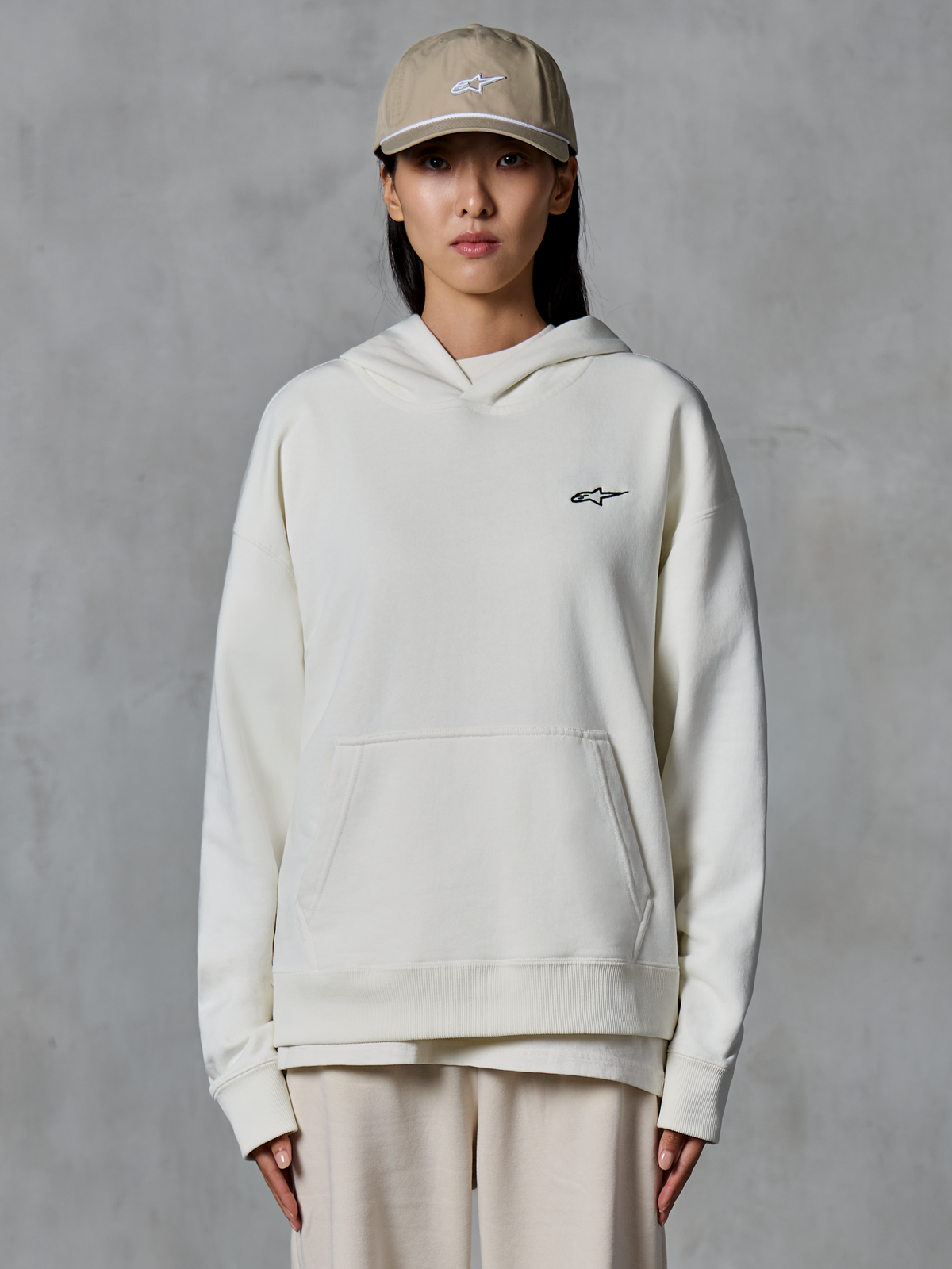 Alpinestars Essentials Ageless Hoodie, felpa con cappuccio in pile color bianco sporco, modella che indossa un cappellino color sabbia e joggers color crema coordinati, piccolo logo Alpinestars nero sul petto, tasca a marsupio frontale, vestibilità rilassata, streetwear casual ispirato al motorsport