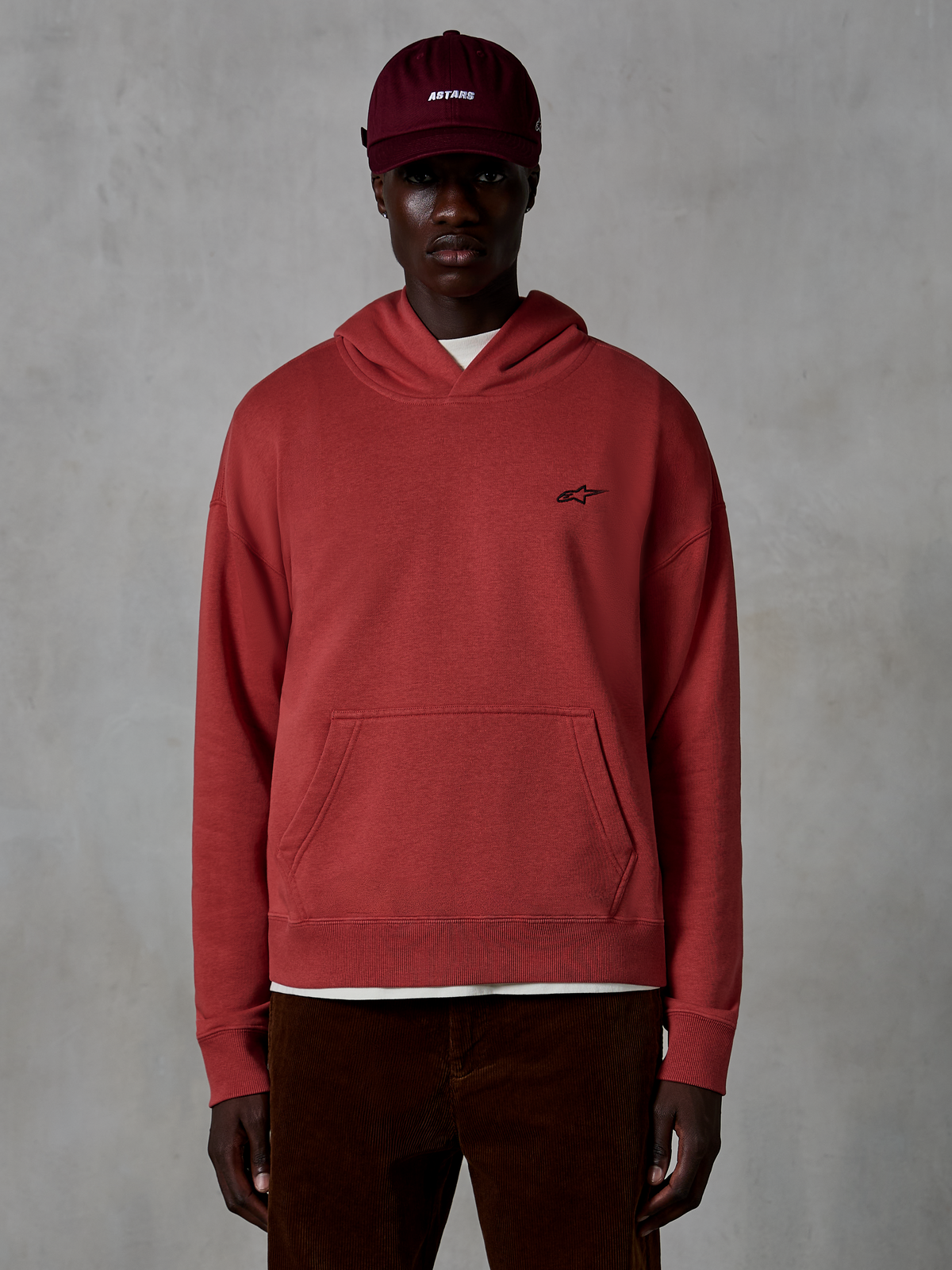Alpinestars Essentials Ageless Hoodie, streetwear casual, colore rosso mattone, con piccolo logo a stella nero sul petto, tasca a marsupio, finitura stone-wash, indossata da un modello con cappellino bordeaux e pantaloni di velluto a coste marroni