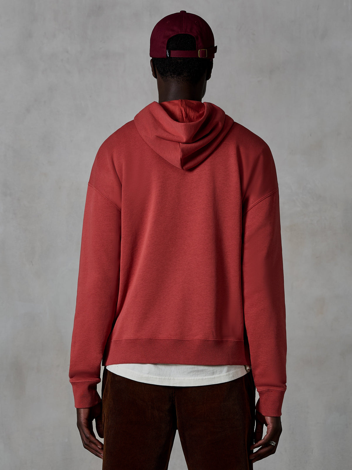 Alpinestars Essentials Ageless Hoodie, felpa casual in pile, rosso mattone, vista posteriore del modello che indossa abbigliamento lifestyle, caratterizzata da un design pulito con orlo e polsini a costine, abbinata a pantaloni in velluto a coste marroni e un berretto bordeaux