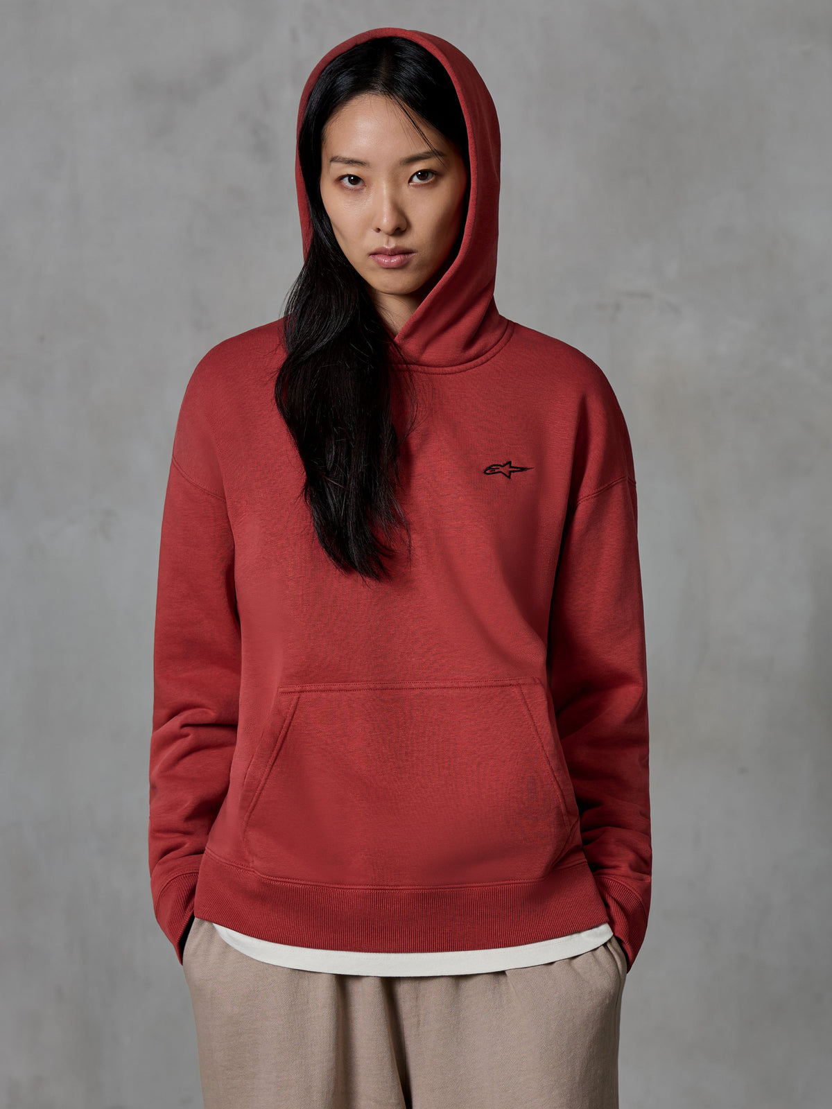 Alpinestars Essentials Ageless Hoodie, felpa pullover rosso mattone, indossata da una modella con il cappuccio alzato, con un piccolo logo a stella nero ricamato sul petto, tasca a marsupio, streetwear casual ispirato al motorsport