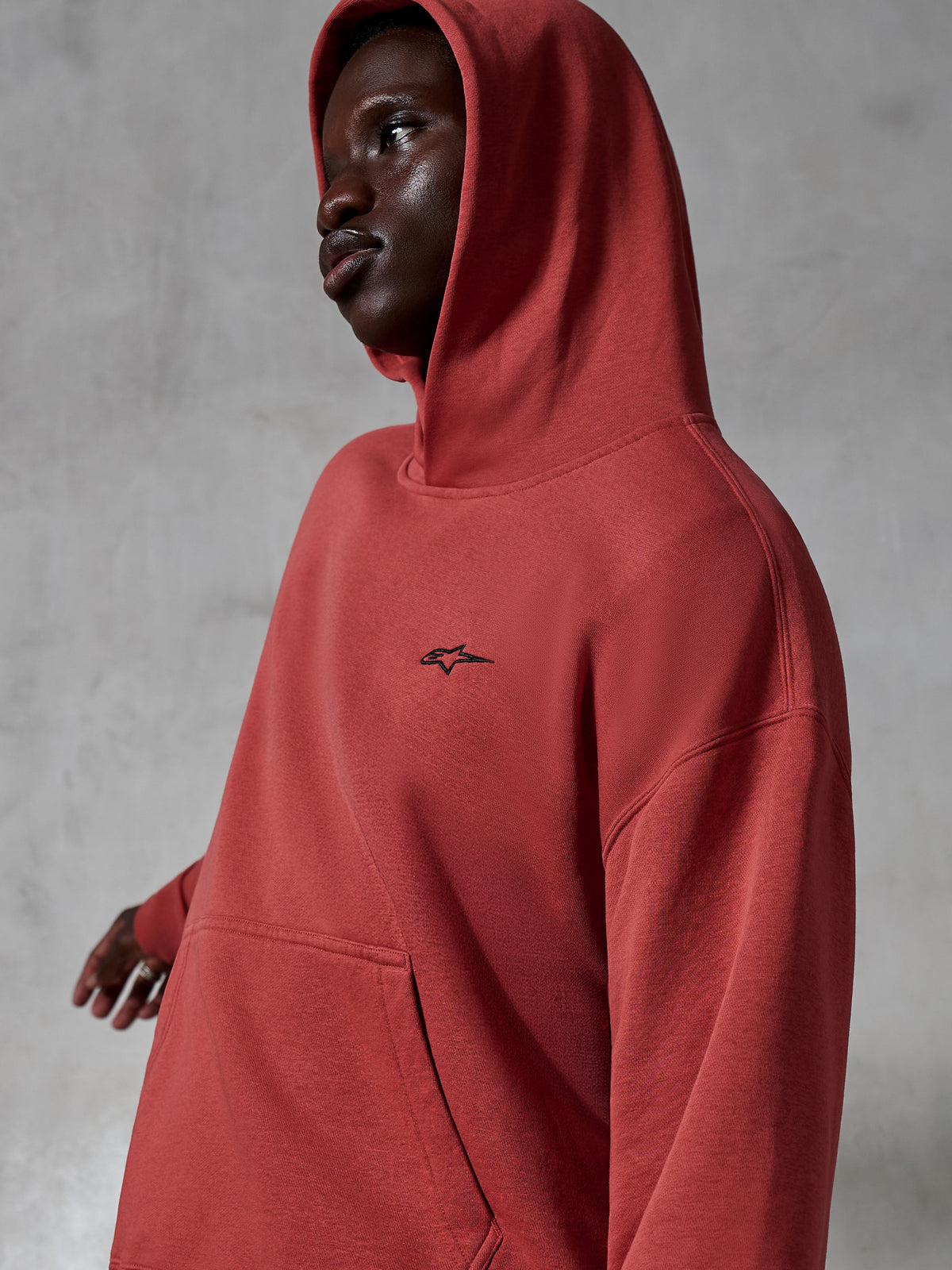 Alpinestars Essentials Ageless Hoodie, abbigliamento sportivo casual, rosso mattone, indossata da un modello con cappuccio sollevato, con un piccolo logo nero ricamato, morbida texture in pile, stile streetwear con vestibilità rilassata