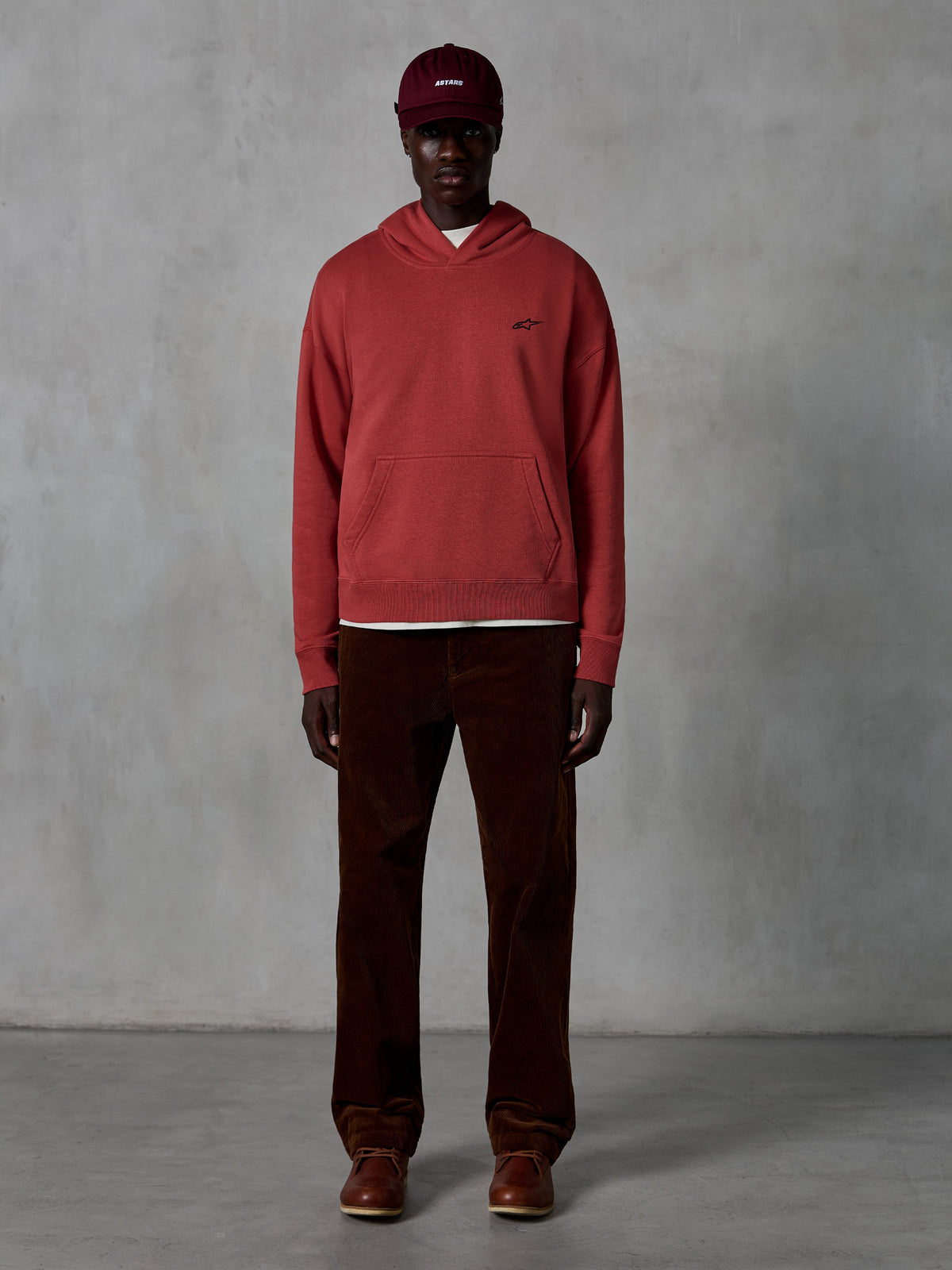 Alpinestars Essentials Ageless Hoodie, felpa casual stile sportswear in colore rosso mattone con finitura stone wash, piccolo logo Alpinestars nero sul petto e tasca a marsupio frontale, indossata da un modello con cappellino bordeaux e pantaloni di velluto a coste marroni per un look streetwear pulito.