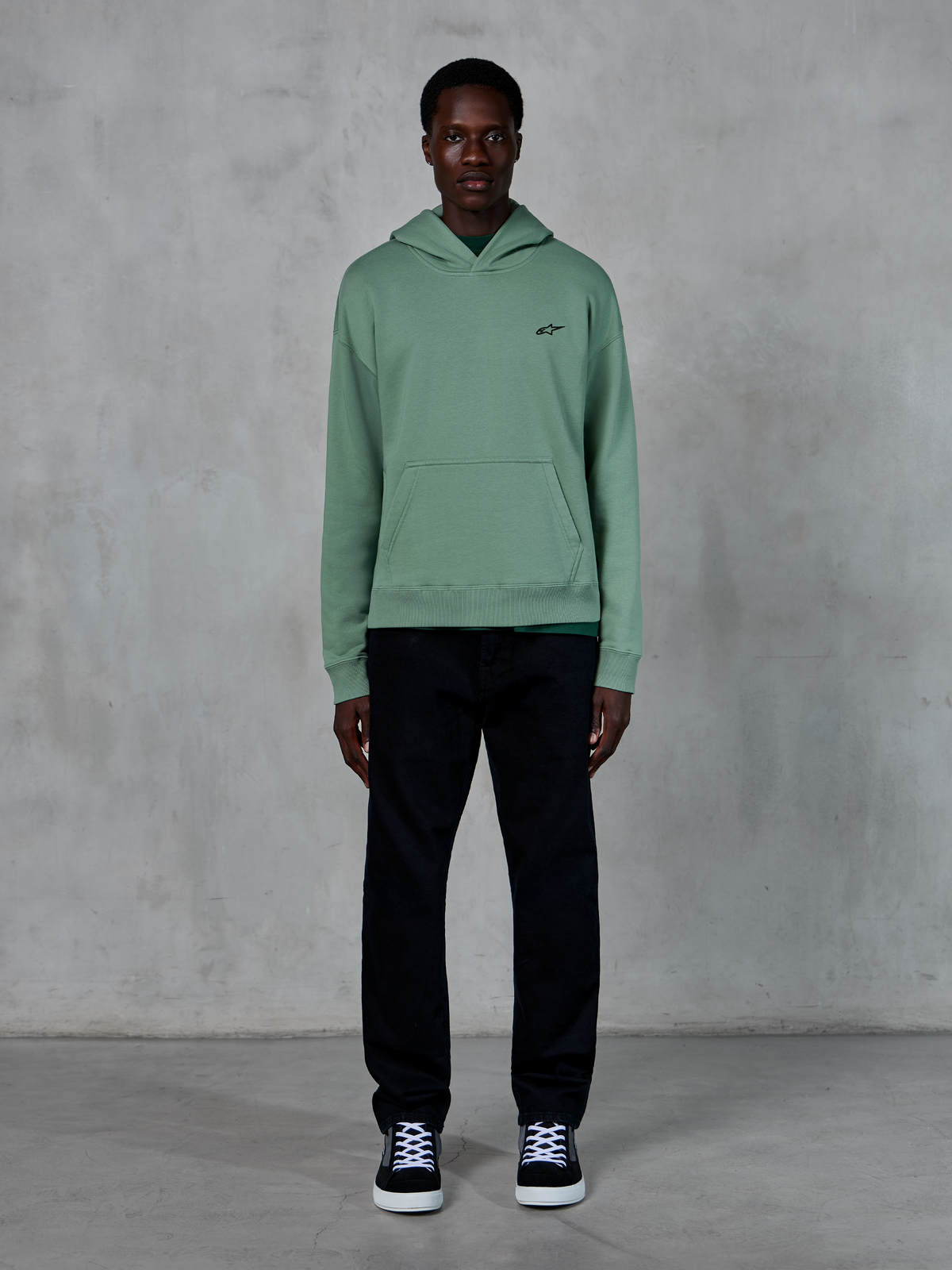 Felpa con cappuccio Alpinestars Essentials Ageless, verde Cactus, piccolo logo nero sul petto, tasca a marsupio, streetwear casual indossato con pantaloni neri e sneaker bianche e nere contro una parete strutturata