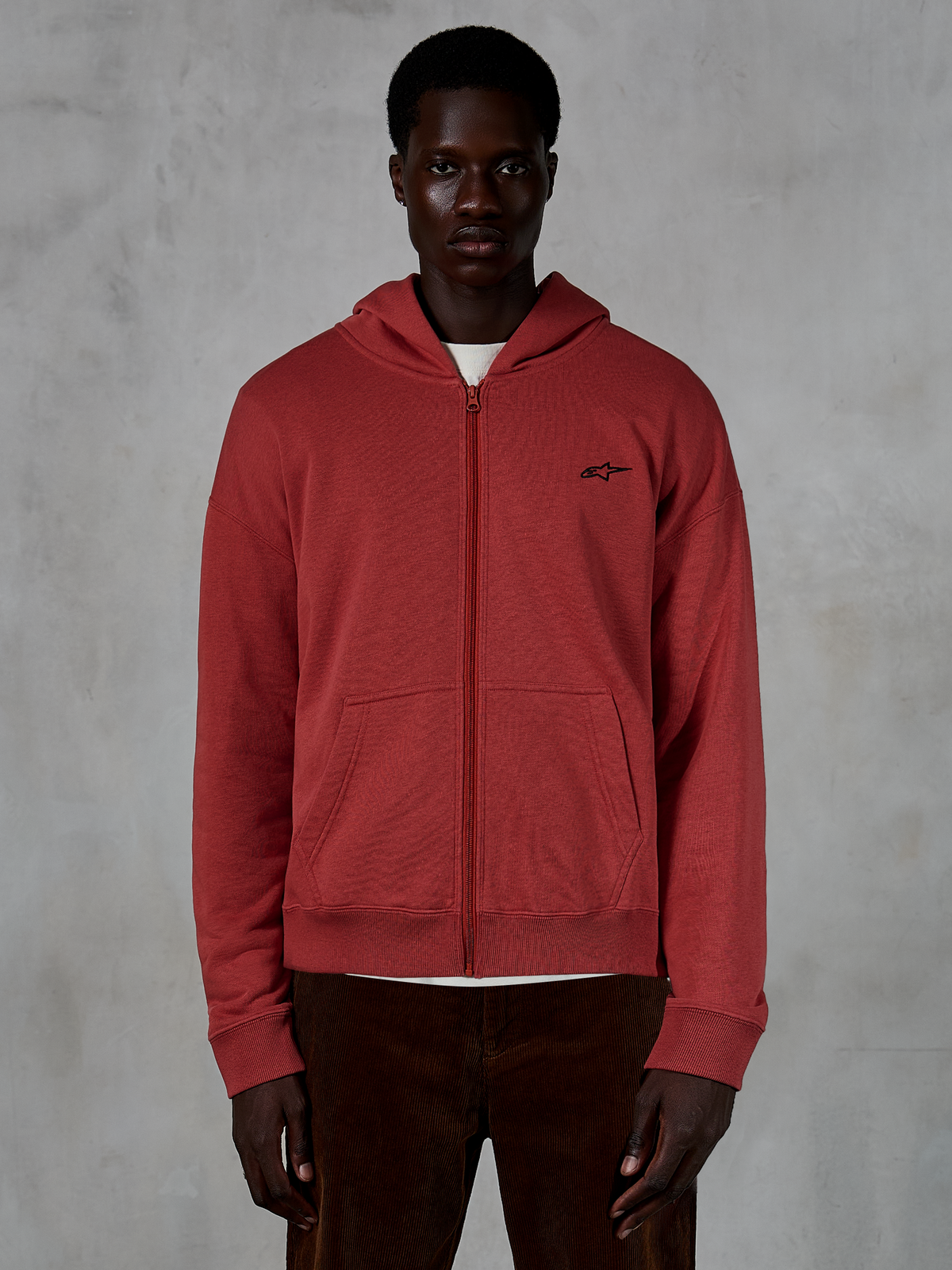 Alpinestars Essentials Ageless Zip Hoodie, felpa con cappuccio e zip integrale rosso mattone, indossata da un modello con pantaloni in velluto a coste marroni, con un piccolo logo nero sul petto, streetwear casual dalla vestibilità rilassata