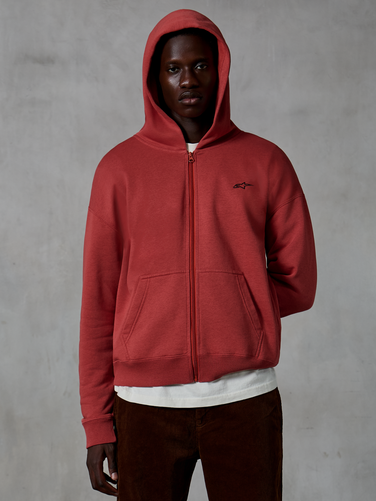 Alpinestars Essentials Ageless Zip Hoodie, abbigliamento sportivo casual in rosso mattone (Brick), design con zip integrale e cappuccio sollevato, piccolo logo nero sul petto, indossata da un modello con t-shirt bianca e pantaloni in velluto a coste marroni su sfondo grigio.