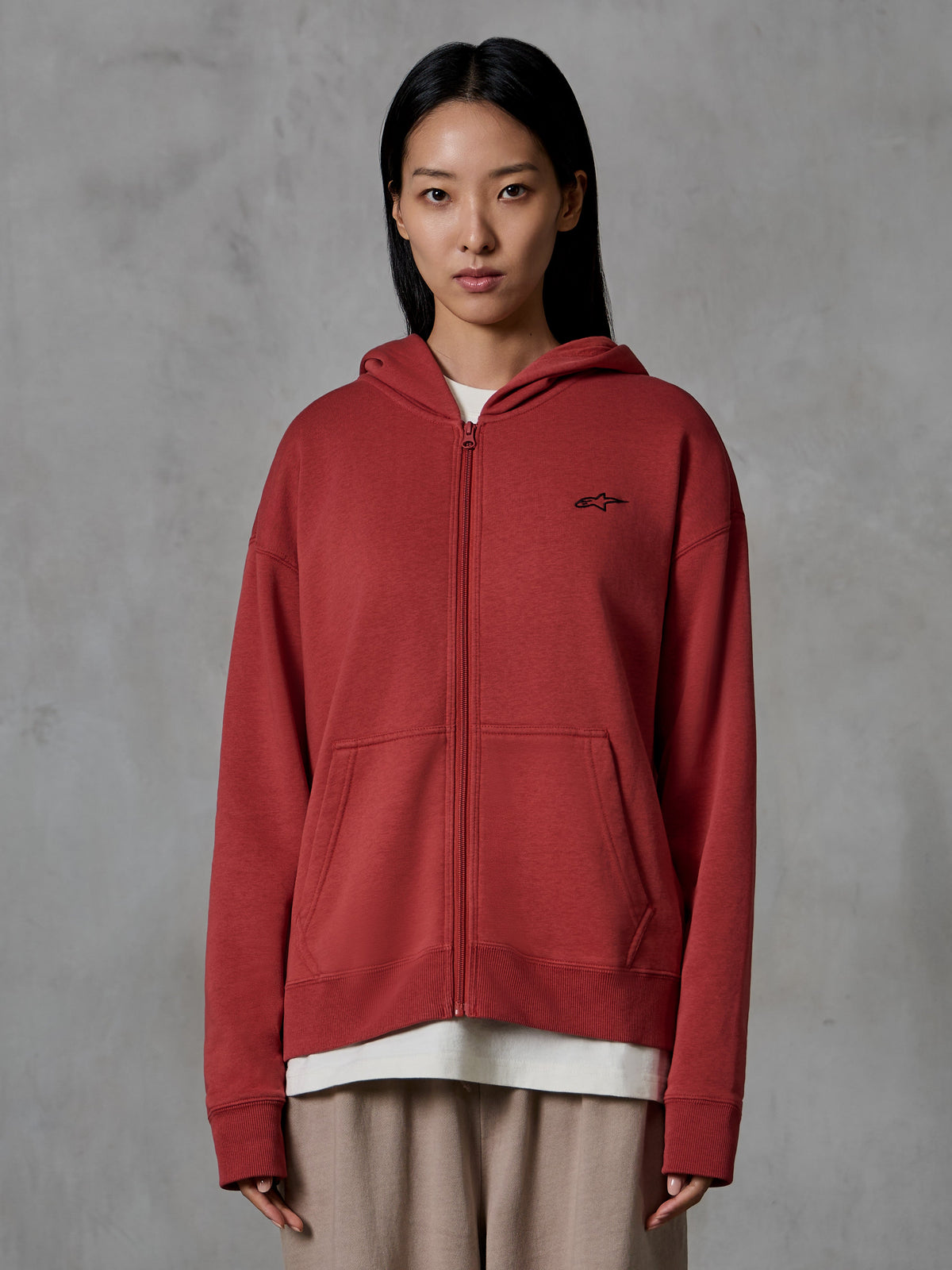 Alpinestars Essentials Ageless Zip Hoodie, rosso mattone, streetwear casual, indossata da una modella, design con zip integrale e cappuccio, piccolo logo Alpinestars nero sul petto, pile compatto dalla vestibilità rilassata.