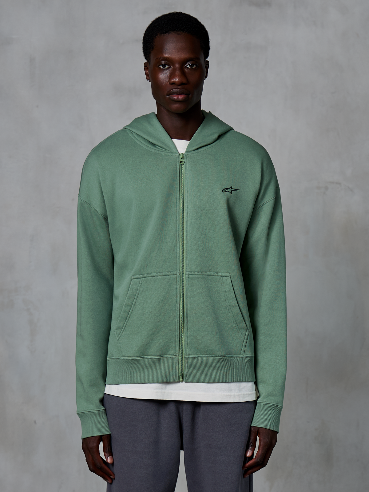 Alpinestars Essentials Ageless Zip Hoodie, felpa casual streetwear con zip, verde Cactus, con un discreto logo ricamato sul petto, tasche a marsupio divise e polsini a costine, indossata da un modello su uno sfondo grigio testurizzato