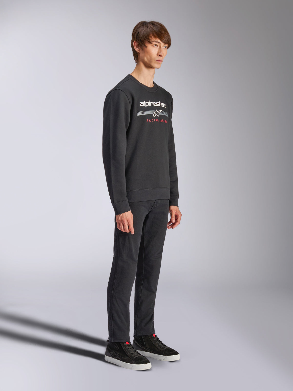 Felpa Alpinestars Bettern Crew, pullover nero a girocollo, logo Alpinestars bianco con scritta 'RACING AHEAD' in rosso sul petto, abbinata a pantaloni neri e sneakers, streetwear casual ispirato al motorsport