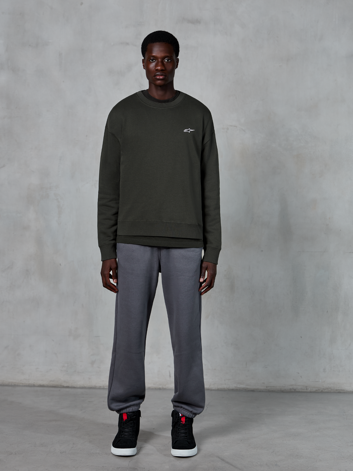 Alpinestars Essentials Ageless Crew, felpa girocollo verde scuro con un piccolo logo bianco sul petto, indossata da un modello con pantaloni della tuta grigi e sneakers alte nere, abbigliamento lifestyle casual
