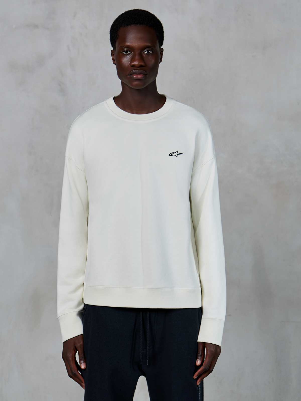 Alpinestars Essentials Ageless Crew, felpa girocollo Off White, piccolo logo Astars nero sul petto a sinistra, abbigliamento sportivo casual lifestyle, tessuto in pile premium, indossata da un modello con joggers neri