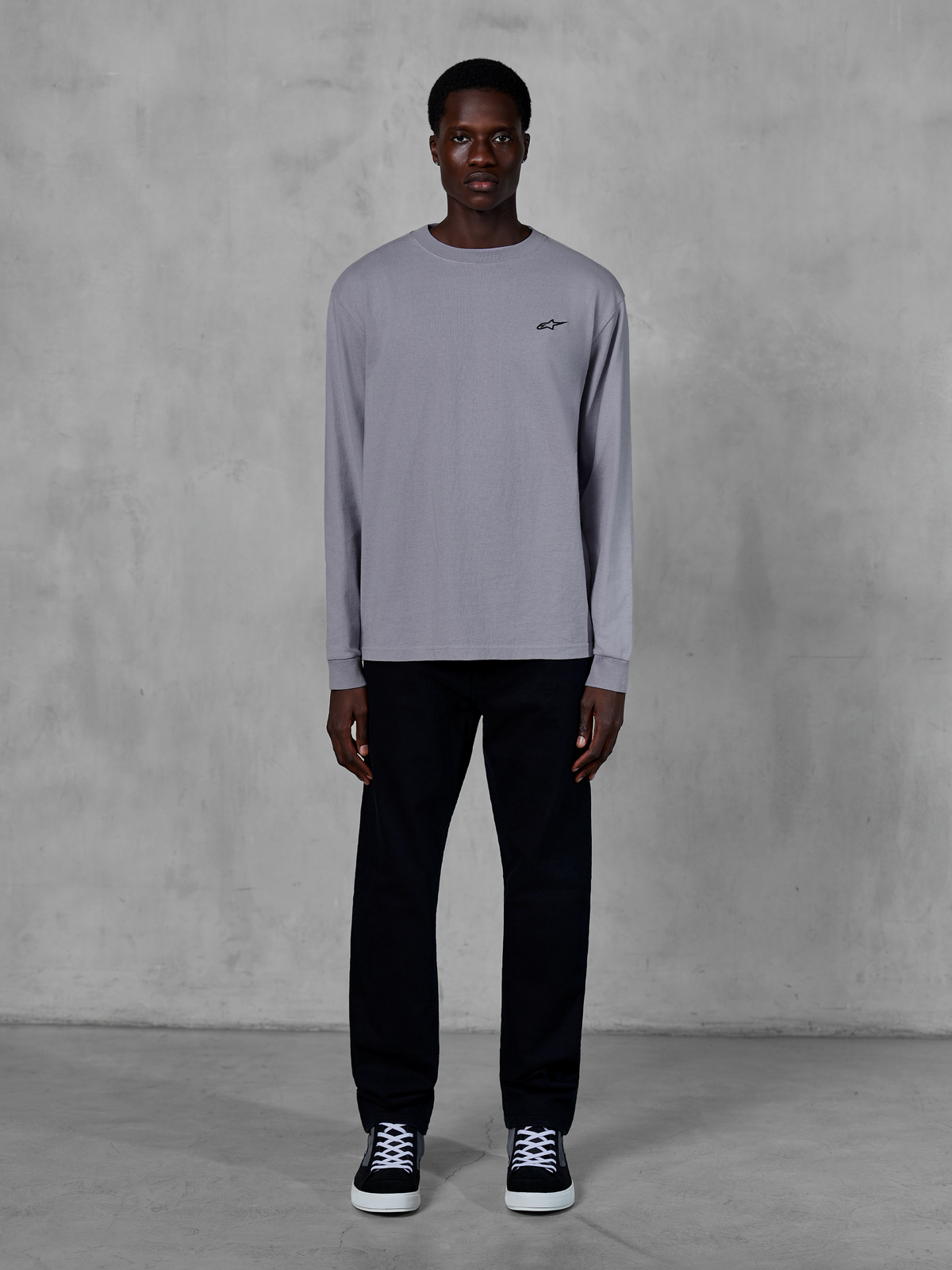Alpinestars Essentials Ageless LS Tee, maglia a maniche lunghe, color argento, piccolo logo Alpinestars nero sul petto, jersey di cotone organico con finitura stone wash, indossata da un modello con pantaloni neri e sneakers, streetwear casual