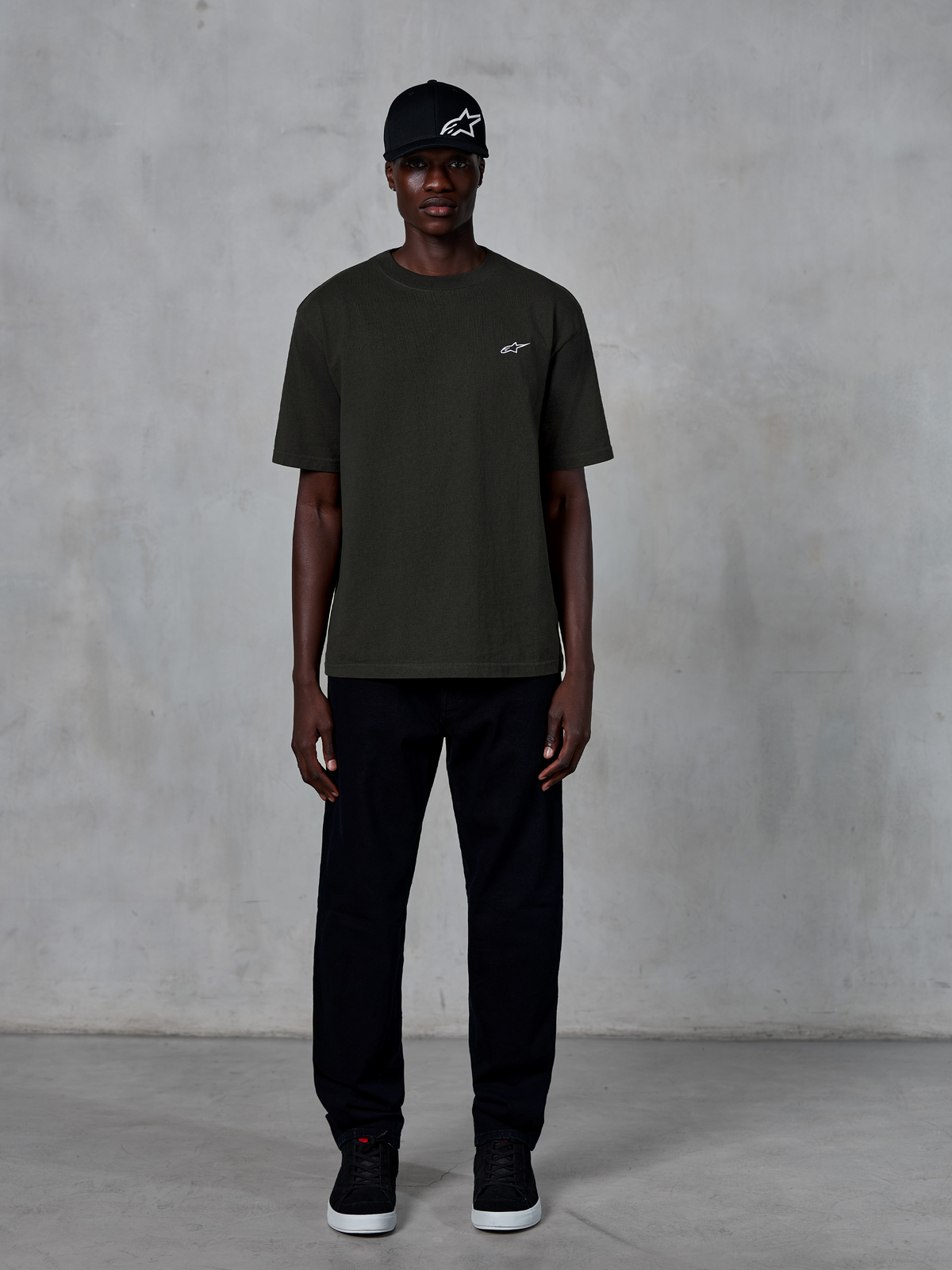 Alpinestars Essentials Ageless SS Tee, maglietta a maniche corte, nero, con un piccolo logo Alpinestars bianco sul petto, indossata da un modello con cappellino e pantaloni neri per un look lifestyle pulito