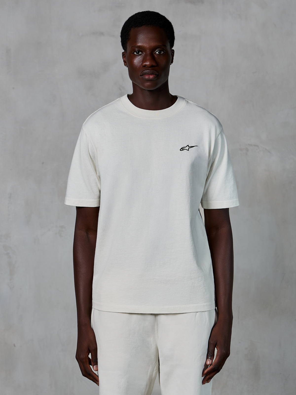 Alpinestars Essentials Ageless SS Tee, t-shirt a maniche corte, off white, indossata da un modello, con un piccolo logo Alpinestars nero sul petto, vestibilità rilassata, jersey di cotone organico, streetwear casual motorsport
