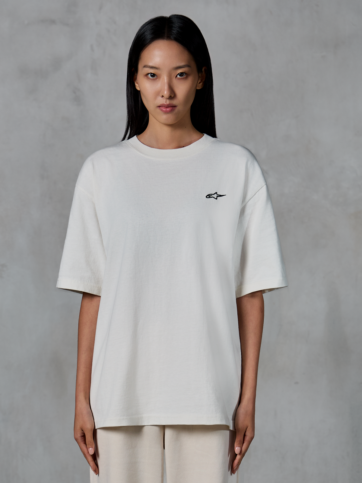 Alpinestars Essentials Ageless SS Tee, maglietta a maniche corte, Off White, indossata da una modella, con un piccolo logo Alpinestars nero sul petto, vestibilità rilassata, abbigliamento casual lifestyle streetwear