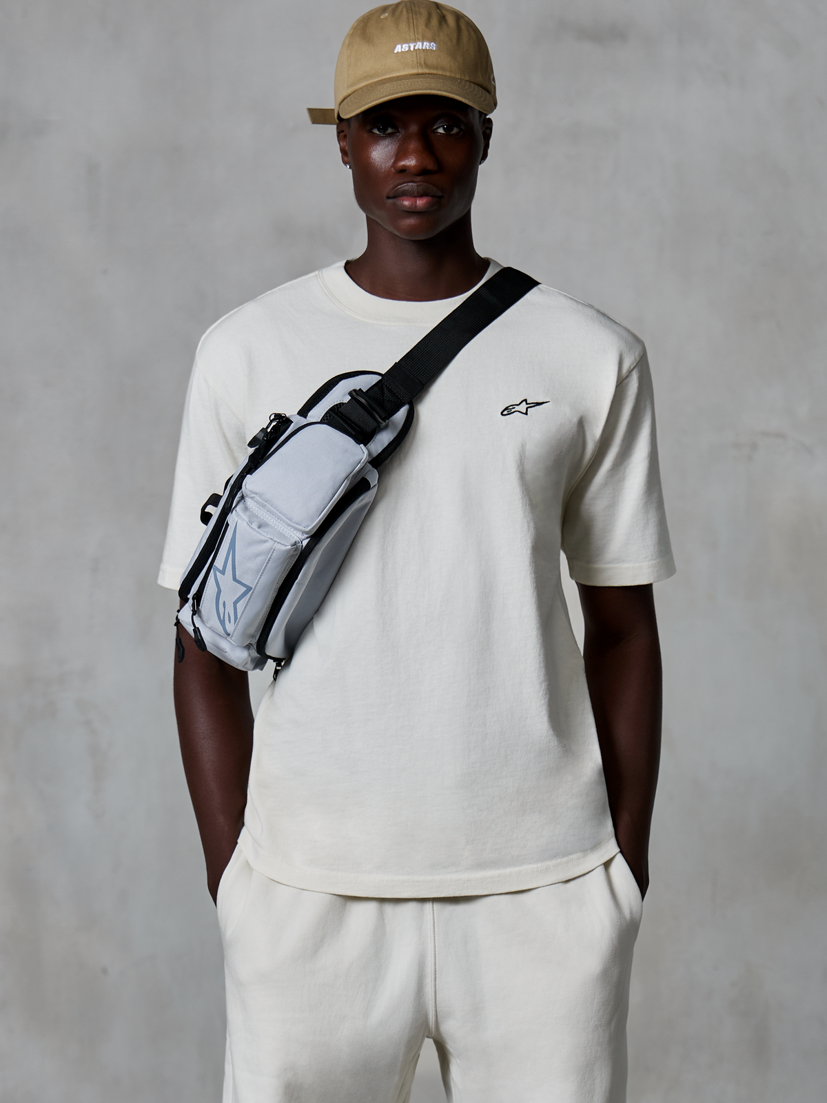 Alpinestars Essentials Ageless SS Tee, t-shirt a maniche corte, Off White, indossata con pantaloncini coordinati e un cappellino beige, con un piccolo logo nero sul petto e una borsa a tracolla grigia, streetwear casual ispirato al motorsport