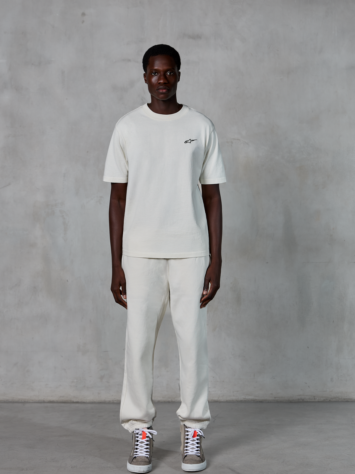 Alpinestars Essentials Ageless SS Tee, maglietta a maniche corte, bianco panna, con un piccolo logo Alpinestars nero sul petto, abbinata a pantaloni sportivi coordinati e sneakers per un look lifestyle rilassato