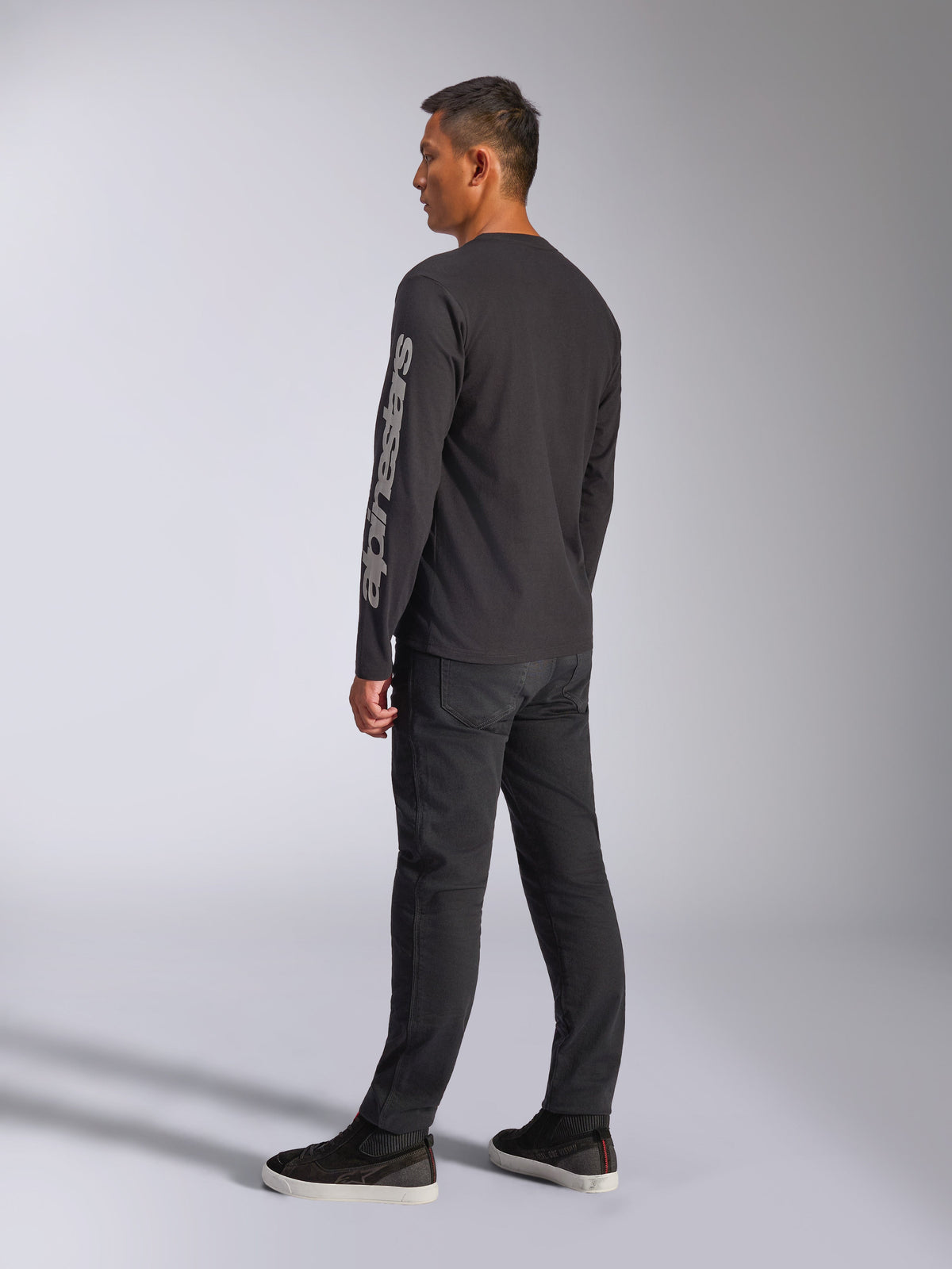 Alpinestars Trackside CSF Long Sleeve Tee, maglia a maniche lunghe nera, con grande logo Alpinestars grigio sulla manica sinistra, indossata da un modello con pantaloni neri e sneakers casual, streetwear casual ispirato al motorsport