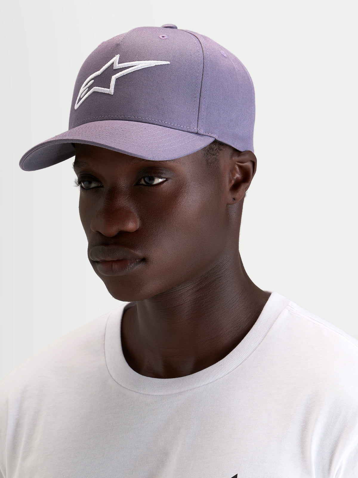 Alpinestars Cappello Ageless Snapback, cappello sportswear con visiera curva in viola polveroso e bianco, indossato da un modello che mette in risalto la corona strutturata e il logo Astars ricamato in 3D