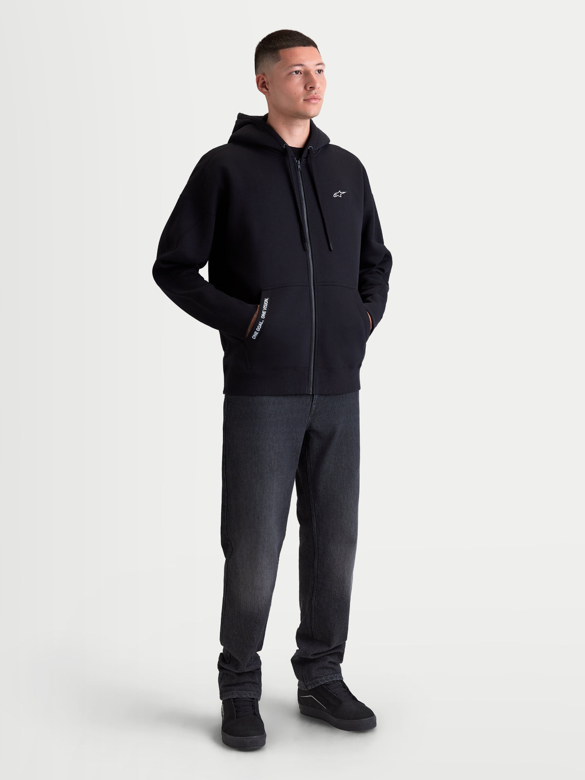 Alpinestars Felpa con cappuccio Pioneer Oversized, felpa da uomo sportswear con zip intera, nera, in misto cotone con cerniera Vislon e tasca a marsupio. Caratterizzata dal logo Astars bianco sul petto e scritta One Goal One Vision. Vista frontale su modello con vestibilità rilassata per abbigliamento casual.