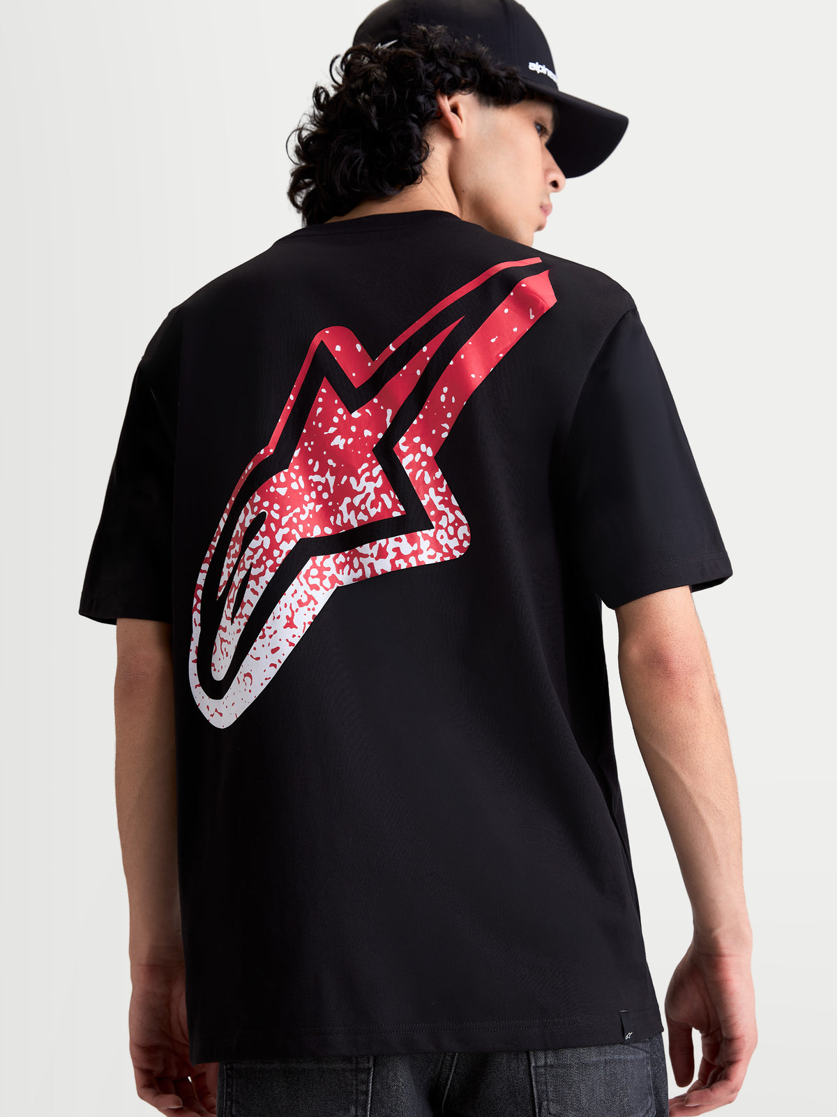 Alpinestars Maglietta Meteor CSF, maglietta a maniche corte nera, vista posteriore su modello con grande logo Astars rosso e bianco sfumato d'ispirazione MX, abbigliamento motorsport casual.