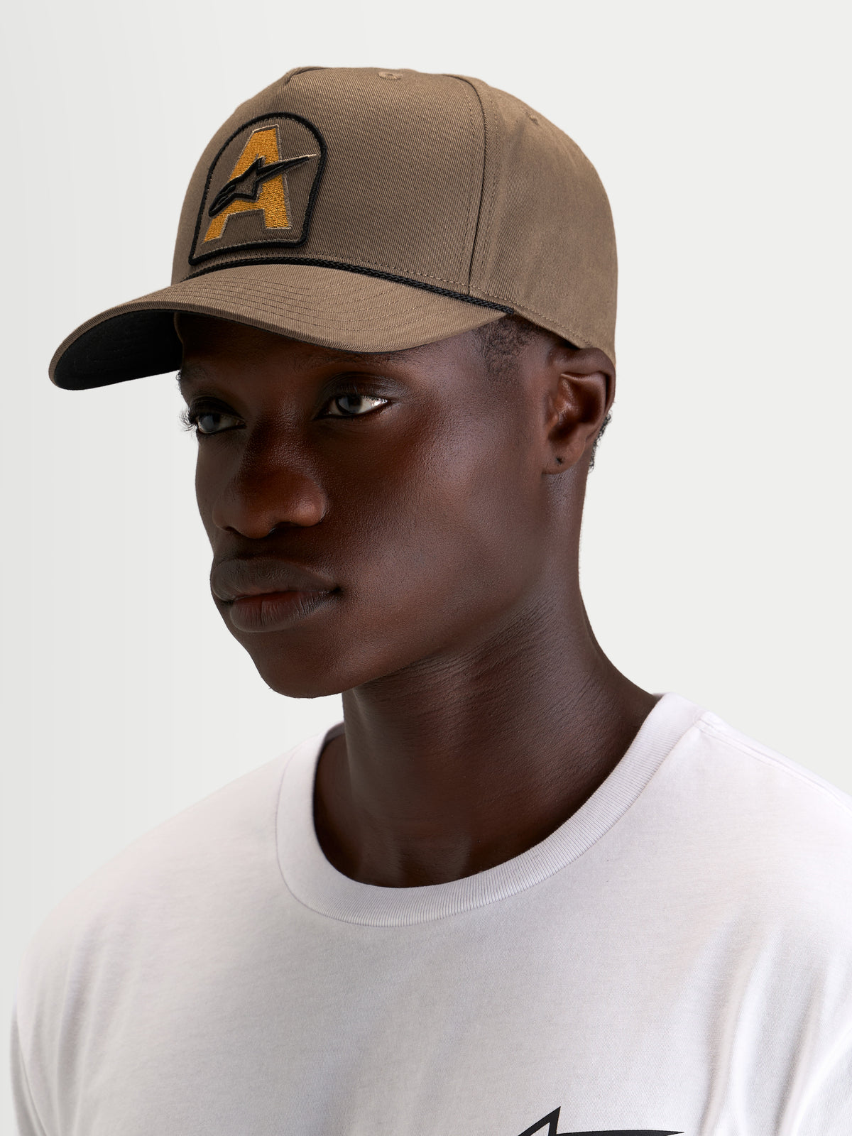 Alpinestars Cappello Acme Snapback, cappello sportswear marrone noce con visiera curva, corona strutturata e patch frontale ricamata del logo Astars con finitura in corda, indossato da un modello