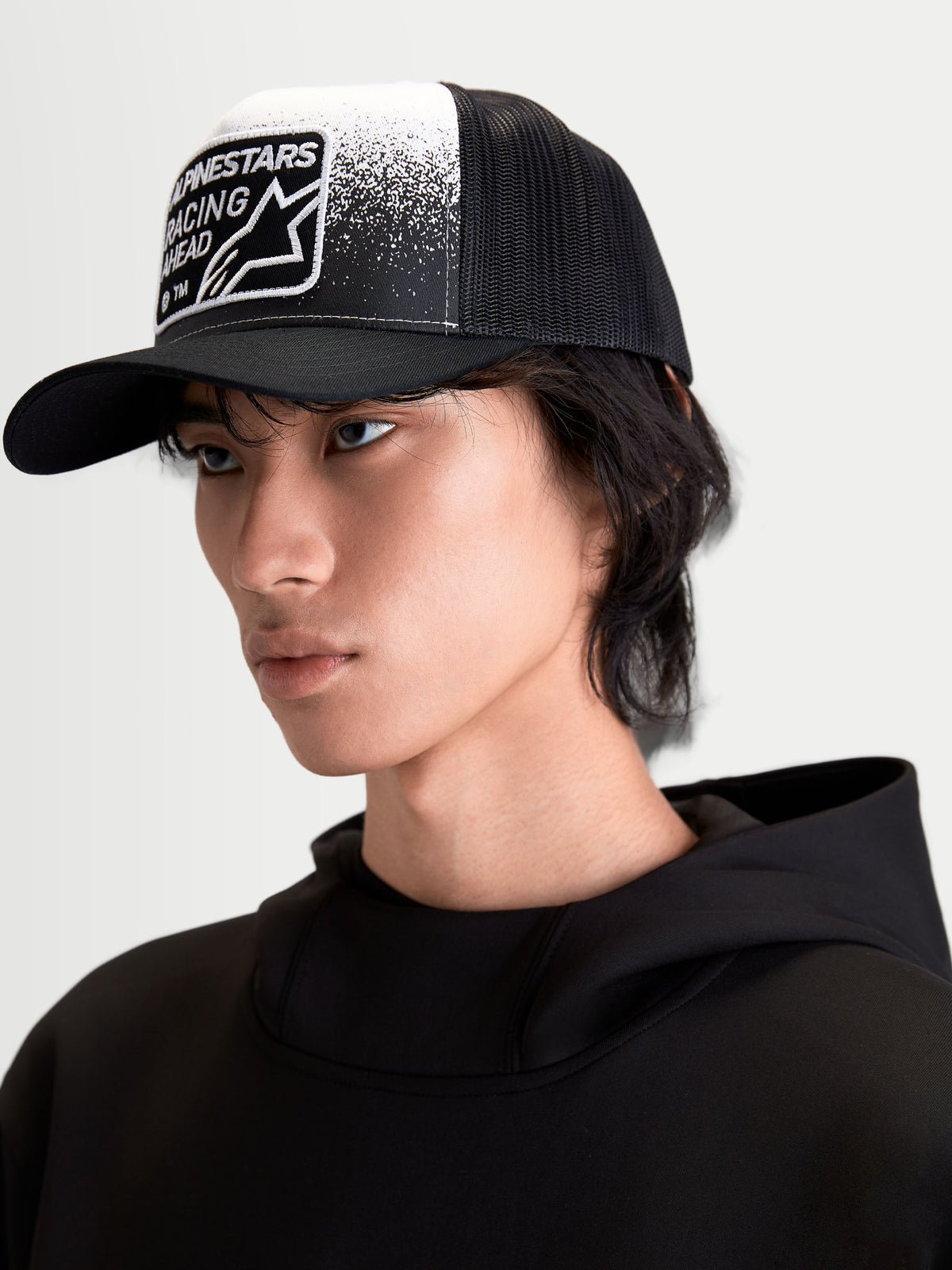 Alpinestars Cappello Gravel, cappello trucker casual indossato da un modello, nero e bianco, con corona strutturata, visiera curva e retro in rete traspirante. Caratterizzato da patch frontale con grafica effetto ghiaia e marchio Alpinestars Racing Ahead.