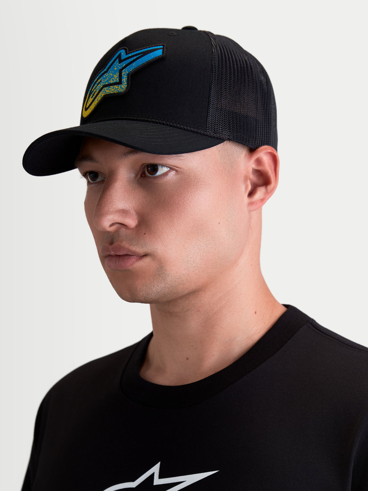Alpinestars Cappello Meteorite, cappello trucker da uomo, nero, patch logo Astars blu e giallo con trama ghiaia, visiera curva, corona strutturata e retro in rete, mostrato su modello.