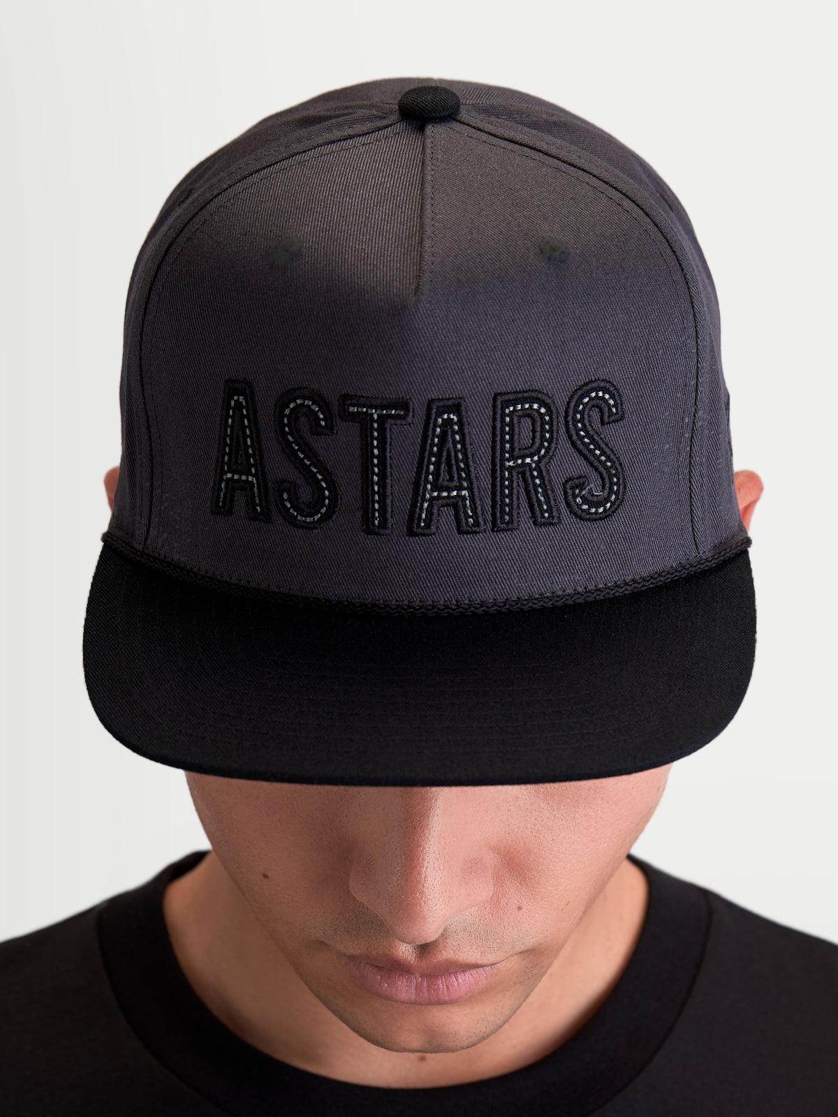 Alpinestars Cappello Stadium Snapback, cappello sportswear unisex ispirato al motorsport, grigio scuro e nero, con visiera piatta, corona strutturata e logo ASTARS ricamato, in twill di cotone, indossato da un modello.