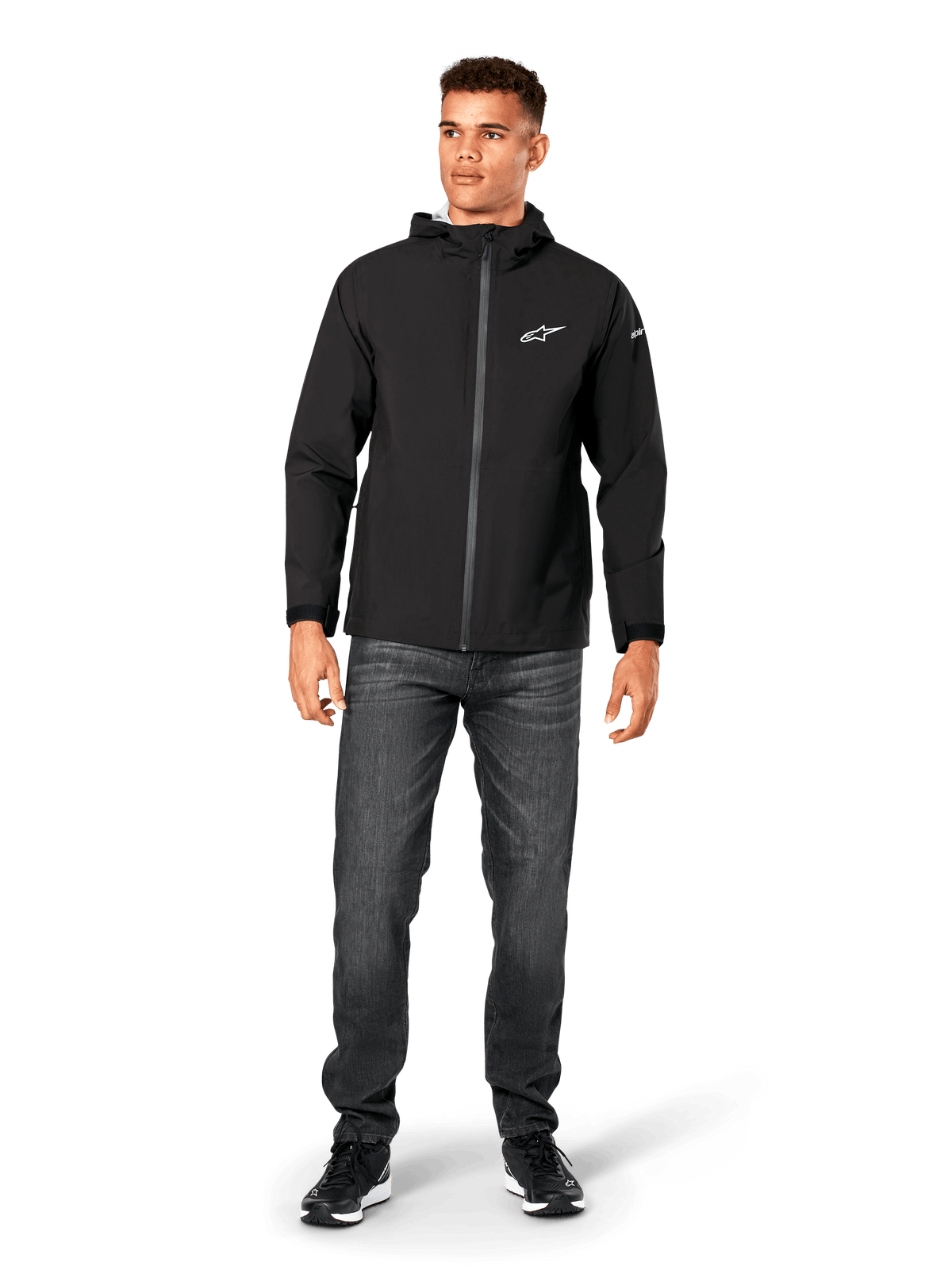 Giacca antipioggia Alpinestars Kascade, shell antipioggia da uomo, nero, indossata con jeans grigio scuro e sneakers nere, caratterizzata da design con cappuccio, cerniera frontale rivestita e logo Alpinestars bianco sul petto, streetwear casual ispirato al motorsport