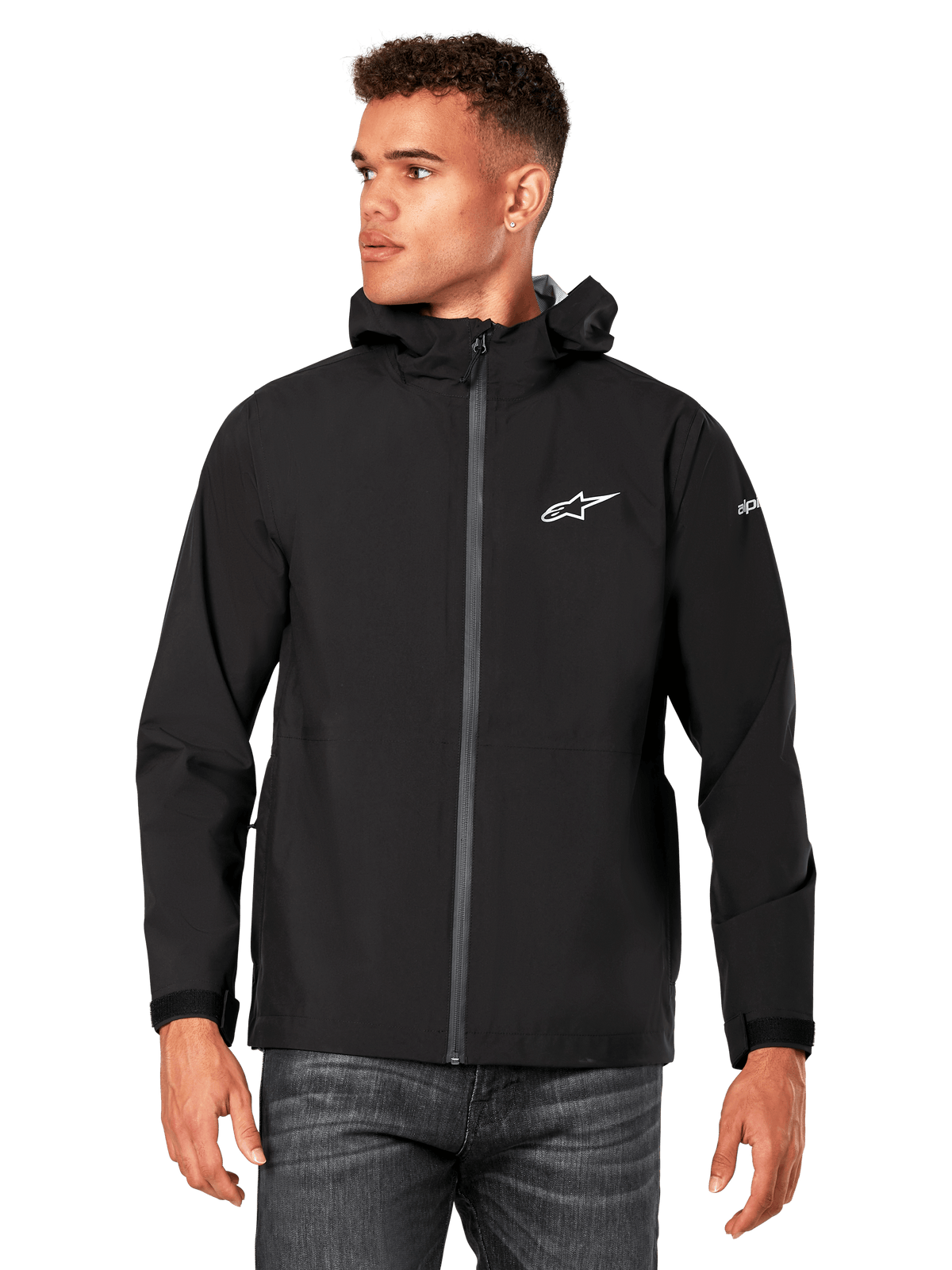 Modello che indossa la giacca antipioggia Alpinestars Kascade, nera, logo bianco sul petto e sulla manica, guscio antipioggia con cappuccio, zip frontale rivestita e polsini in velcro, abbinata a jeans scuri, streetwear casual ispirato al motorsport
