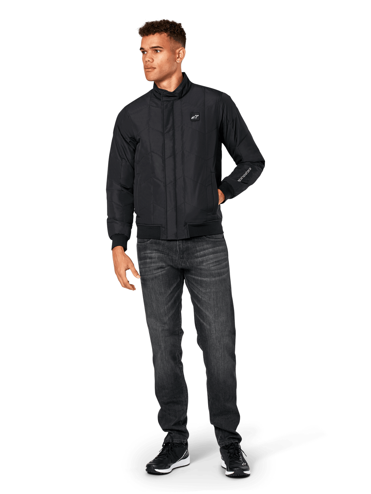 Giacca Alpinestars Precedent, giacca sportswear trapuntata, nera, indossata con jeans grigio scuro e sneakers nere, con motivo trapuntato angolato, colletto basso, polsini e orlo a coste, patch logo bianca sul petto e ricamo sulla manica.