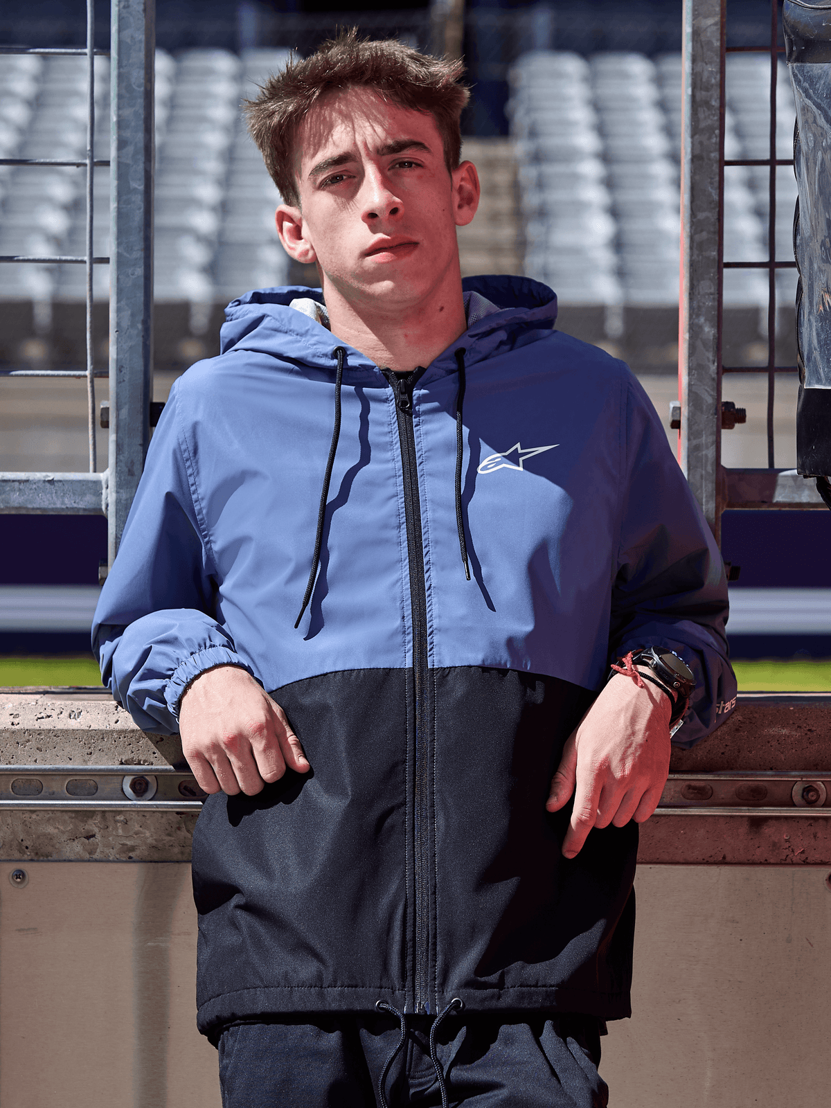 Giacca a vento Alpinestars Speeder, blu ardesia e nero, giacca a vento con cappuccio e design a blocchi, logo bianco sul petto, abbigliamento lifestyle casual indossato in un circuito