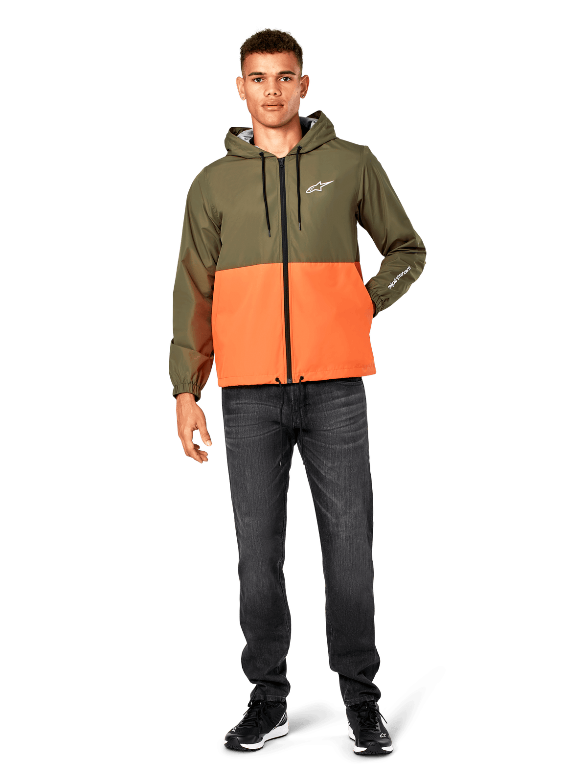 Giacca a vento Alpinestars Speeder, design color-block verde militare e arancione, giacca leggera con cappuccio, cordini neri e zip frontale, logo bianco su petto e manica, indossata con jeans scuri e sneaker nere, stile casual sportswear