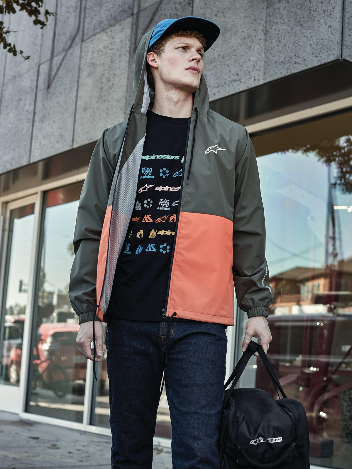 Uomo che indossa la giacca a vento Alpinestars Speeder in Military Orange, con design a blocchi di colore verde militare e arancione, cappuccio con fodera in rete, abbinata a una t-shirt nera con logo grafico, jeans blu e borsone Alpinestars nero, look casual sportswear lifestyle