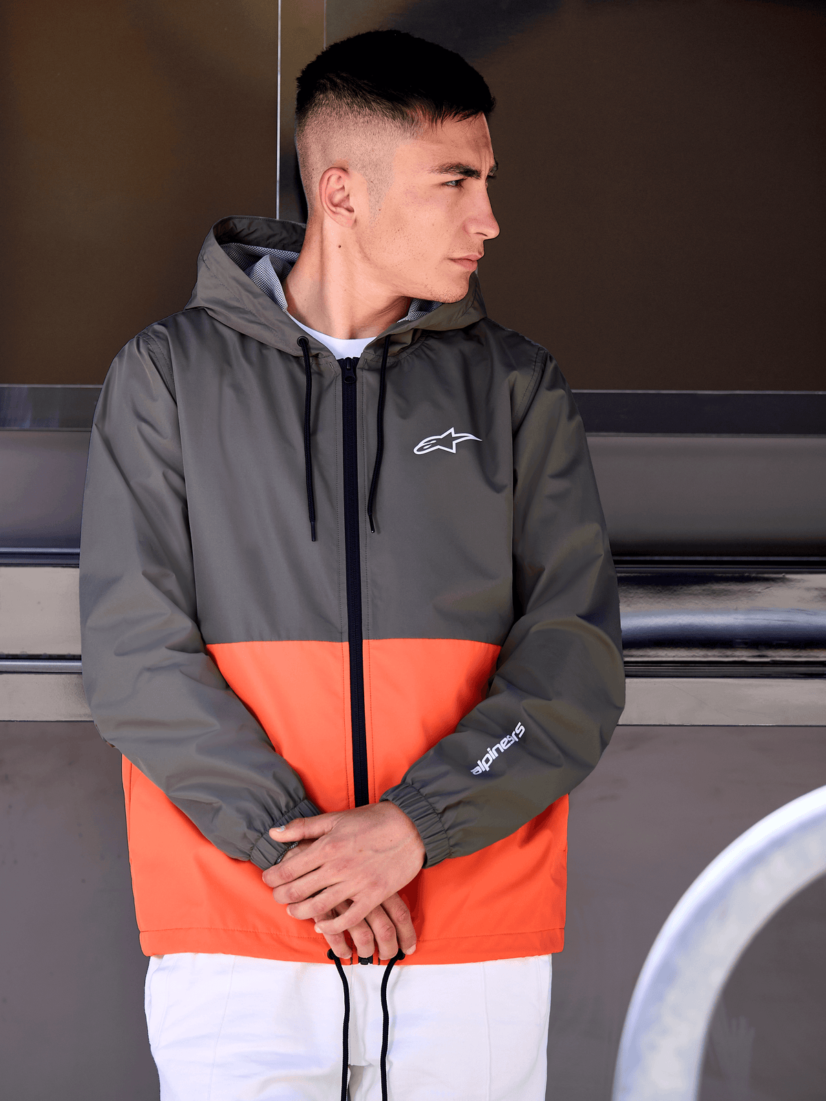 Uomo con giacca a vento Alpinestars Speeder, design a blocchi di colore arancione militare e verde scuro, giacca casual con cappuccio e loghi bianchi su petto e manica, abbinata a pantaloni bianchi per un look lifestyle ispirato al motorsport
