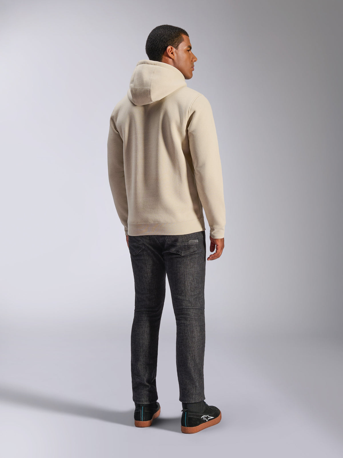 Alpinestars Ageless V3 Hoodie, felpa casual con cappuccio in pile bianco sporco vista dal retro, caratterizzata da polsini e orlo a costine, indossata con jeans scuri slim-fit e sneakers Alpinestars nere