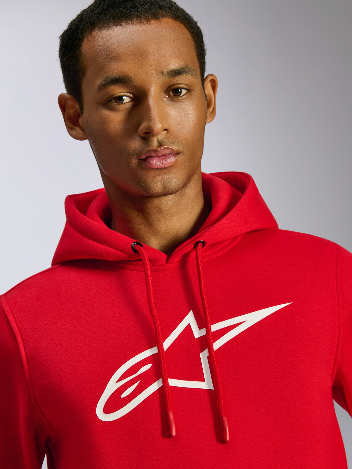 Alpinestars Ageless V3 Hoodie, felpa con cappuccio rossa, grande logo Alpinestars bianco sul petto, cappuccio con coulisse ed estremità immerse, streetwear casual ispirato al motorsport, indossata da un modello uomo