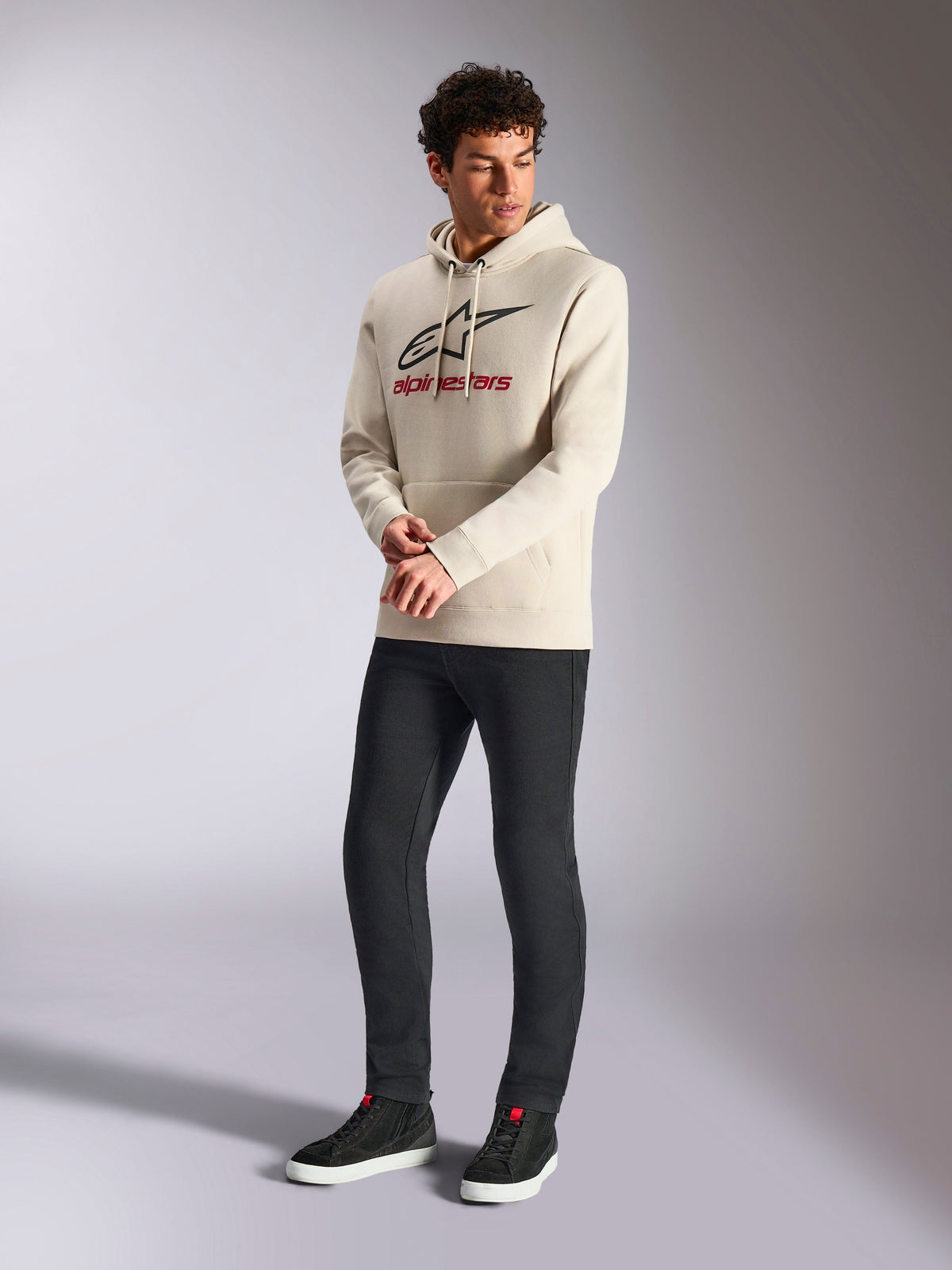 Alpinestars Always V3 Hoodie, felpa in pile con cappuccio bianco sporco, logo lineare frontale con stella nera e marchio rosso, indossata da un modello con pantaloni neri e sneakers nere, streetwear casual motorsport