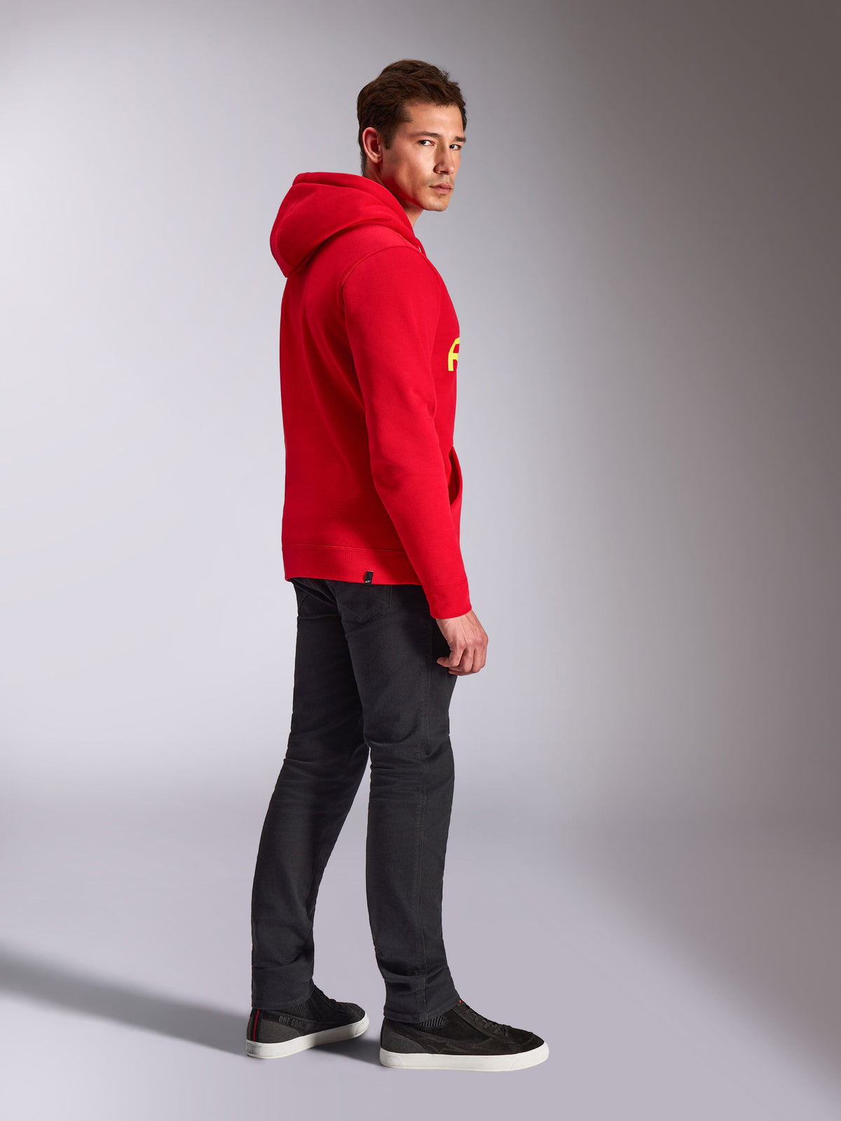 Felpa con cappuccio Alpinestars Linear Wordmark V3, rosso, streetwear casual, modello visto di lato e da dietro che indossa una felpa rossa con marchio giallo, jeans grigio scuro e sneakers nere, scatto lifestyle in studio