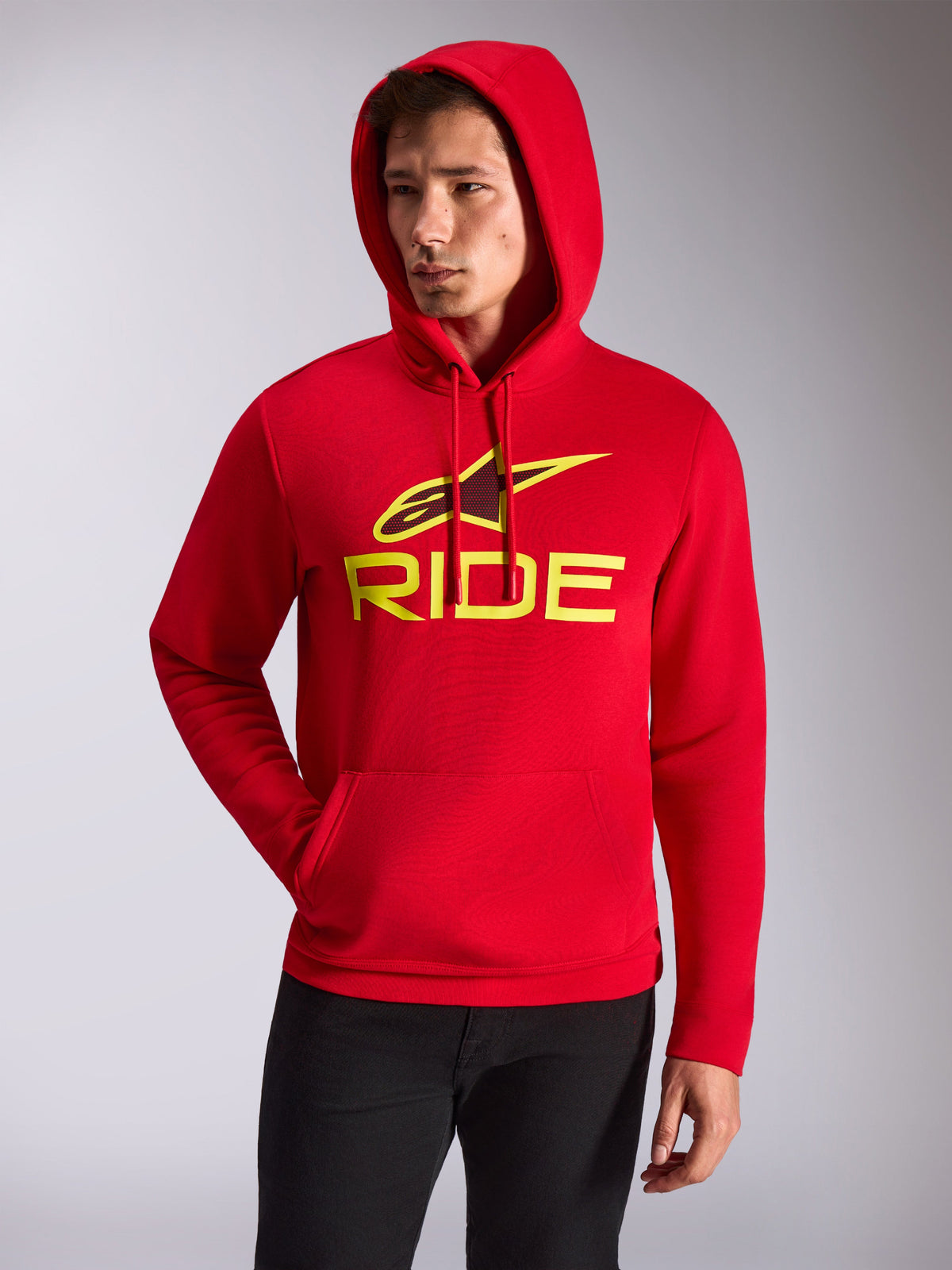 Alpinestars Ride V3 Hoodie, modello maschile che indossa una felpa rossa con cappuccio in pile, logo RIDE giallo fluo e stella Alpinestars sul petto, streetwear casual motorsport, tasca a marsupio anteriore, abbinata a pantaloni neri