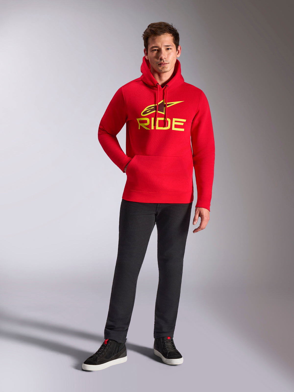 Alpinestars Ride V3 Hoodie, felpa in pile rossa, logo RIDE giallo vivace con simbolo Astars sul petto, indossata con pantaloni neri e sneakers alte nere, abbigliamento streetwear casual ispirato al motorsport