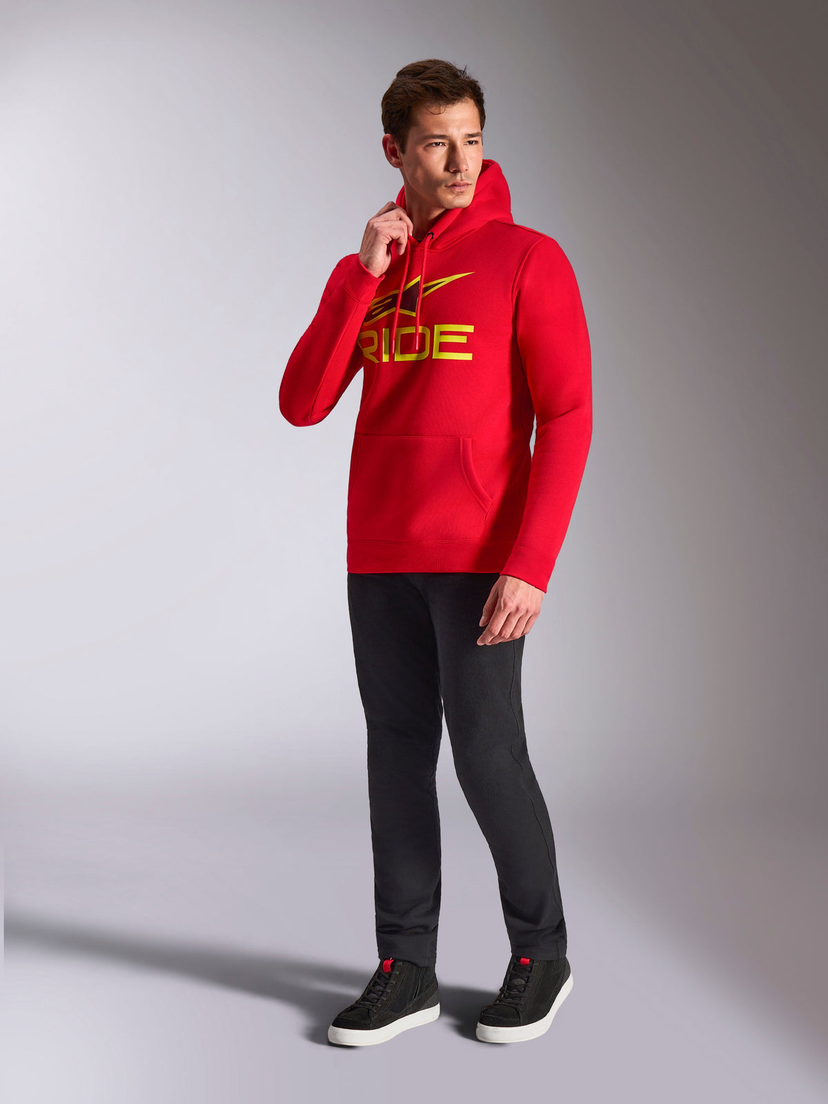 Alpinestars Ride V3 Hoodie, felpa pullover rossa, logo 'RIDE' giallo e nero sul petto, indossata da un modello con pantaloni neri e sneakers nere, streetwear casual ispirato al motorsport