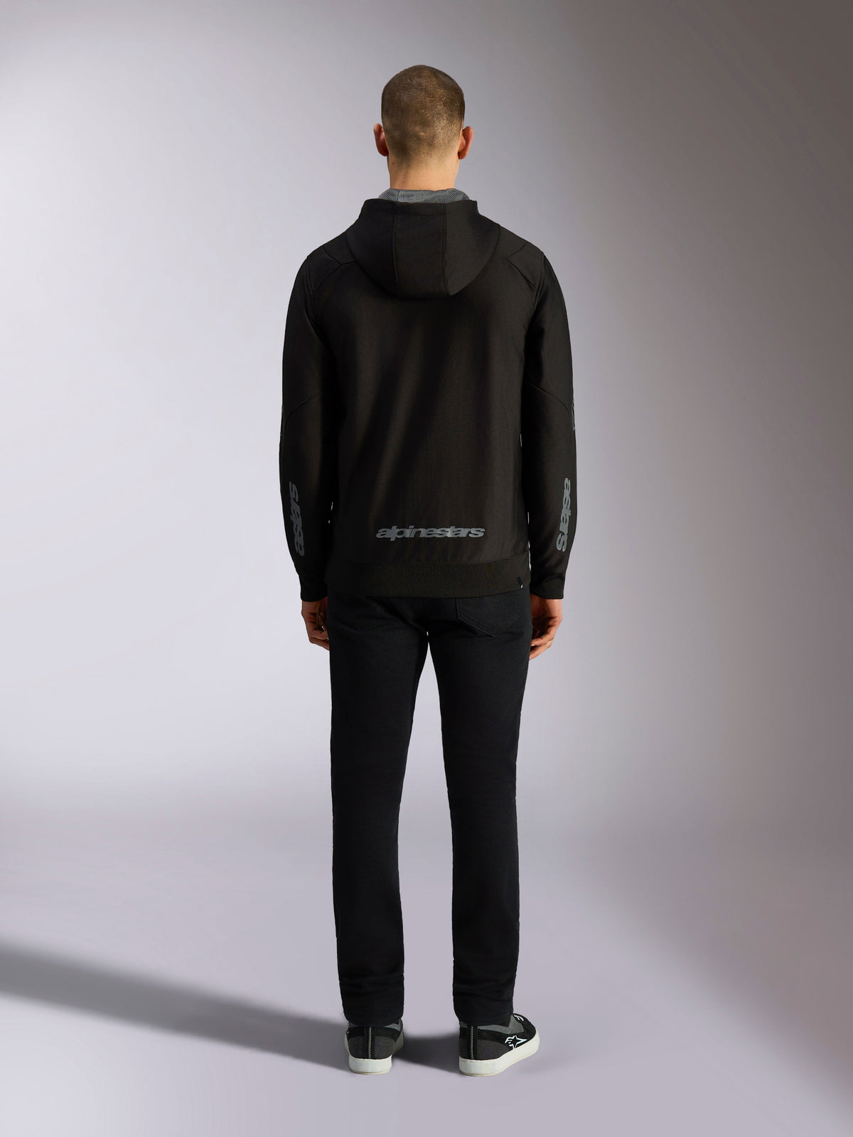 Vista posteriore della felpa con cappuccio Alpinestars Sessions V3 in nero e grigio, con scritta Alpinestars grigia sulla parte inferiore della schiena e loghi astars sugli avambracci, indossata da un modello con pantaloni neri, streetwear casual ispirato al motorsport