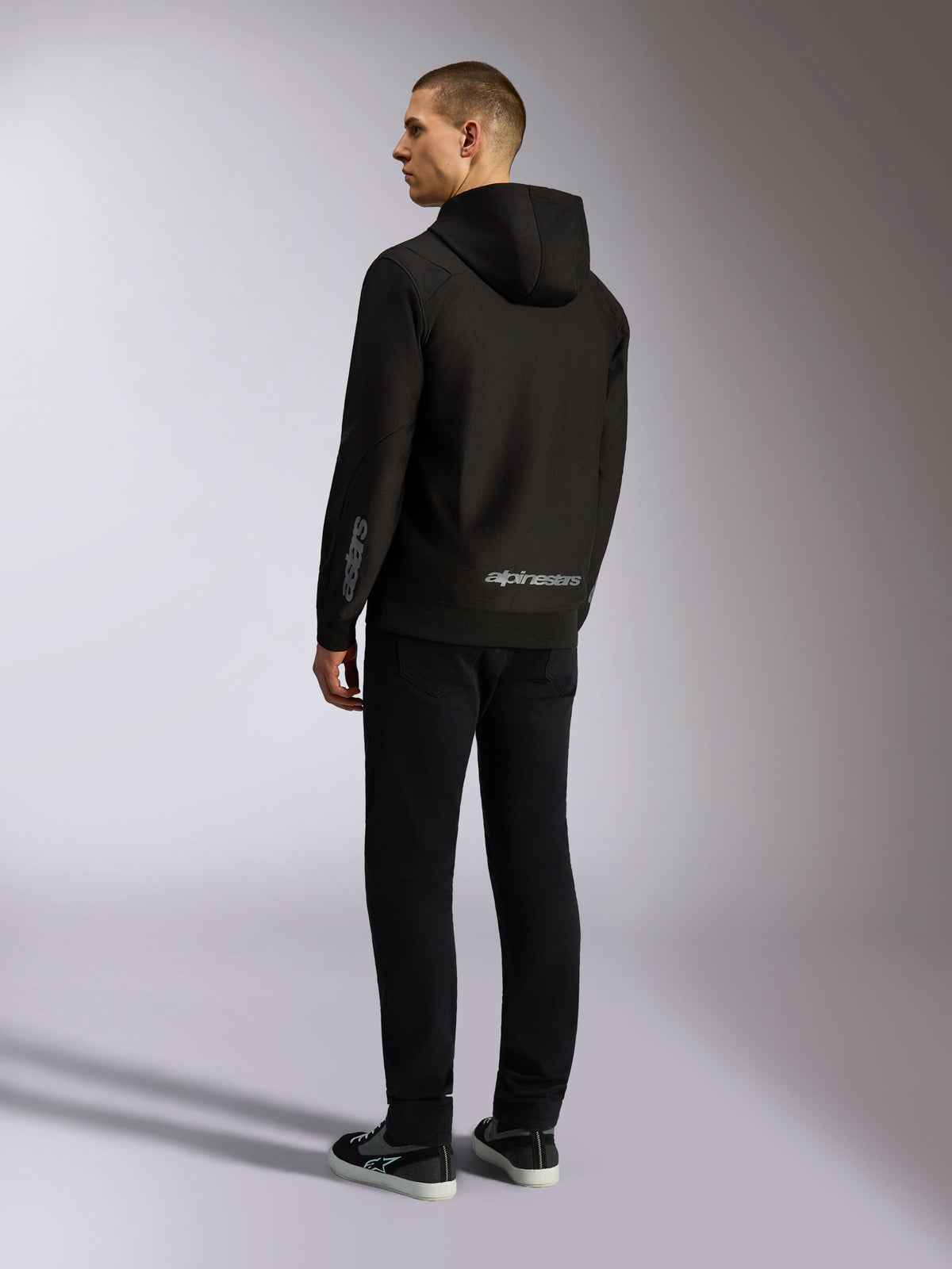 Felpa con cappuccio Alpinestars Sessions V3, streetwear casual nero, vista posteriore su un modello, con scritta Alpinestars grigia sulla parte inferiore della schiena e logo Astars sulla manica, design con cappuccio e zip frontale dallo stile moderno ispirato al motorsport