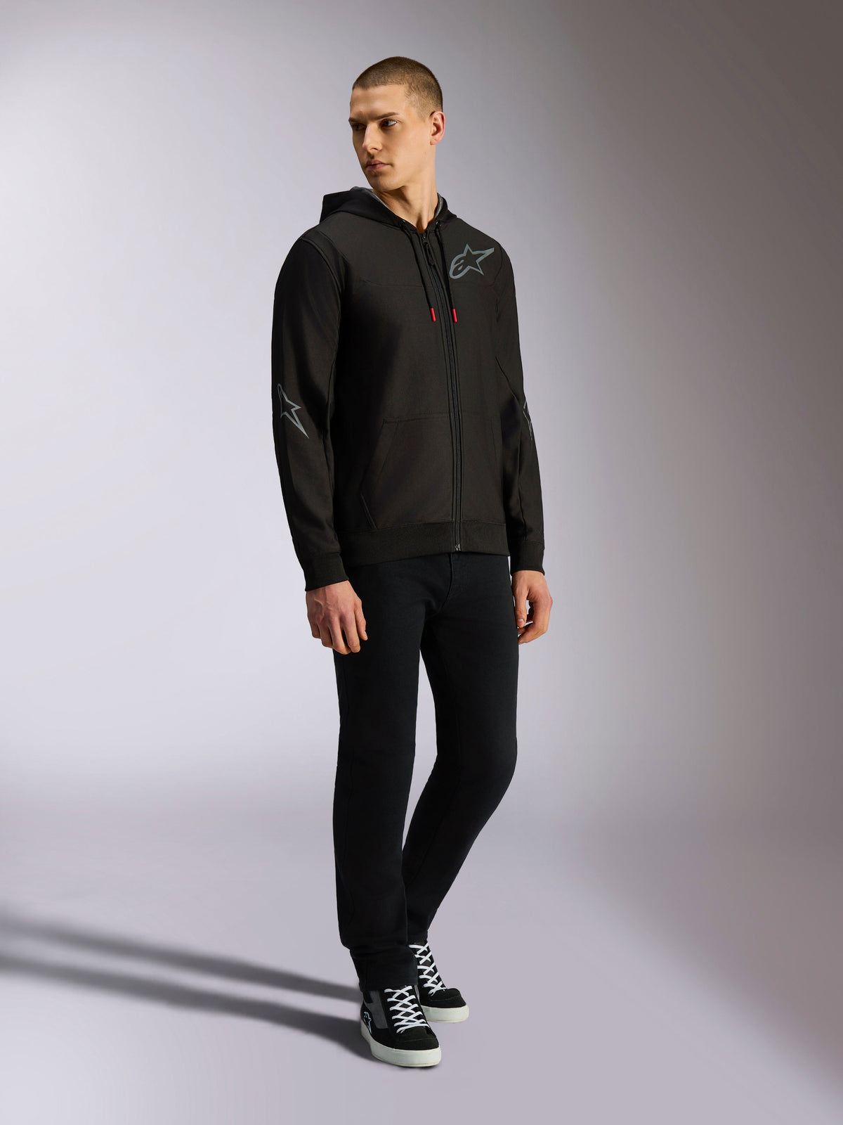Alpinestars Sessions V3 Hoodie, felpa in pile di poliestere con zip frontale, nera e grigia, loghi grigi su petto e maniche, cappuccio con coulisse e punte dei cordini rosse, vista a figura intera del modello in abbigliamento casual motorsport streetwear