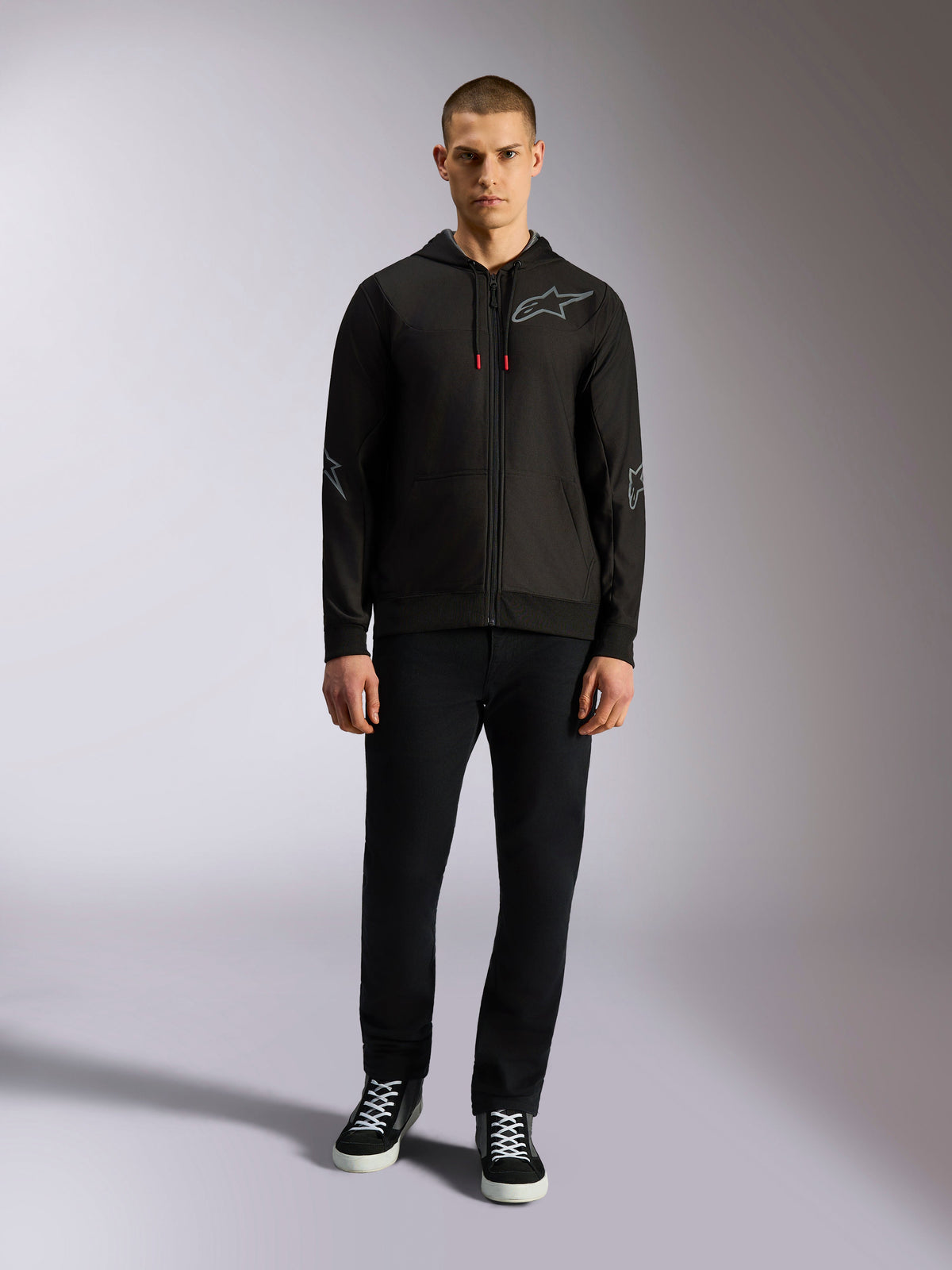 Alpinestars Sessions V3 Hoodie, felpa nera con zip integrale e logo Astars grigio sul petto, grafiche su gomiti e avambracci, dettagli dei cordini in rosso, indossata da modello uomo con pantaloni neri e sneakers, abbigliamento casual motorsport