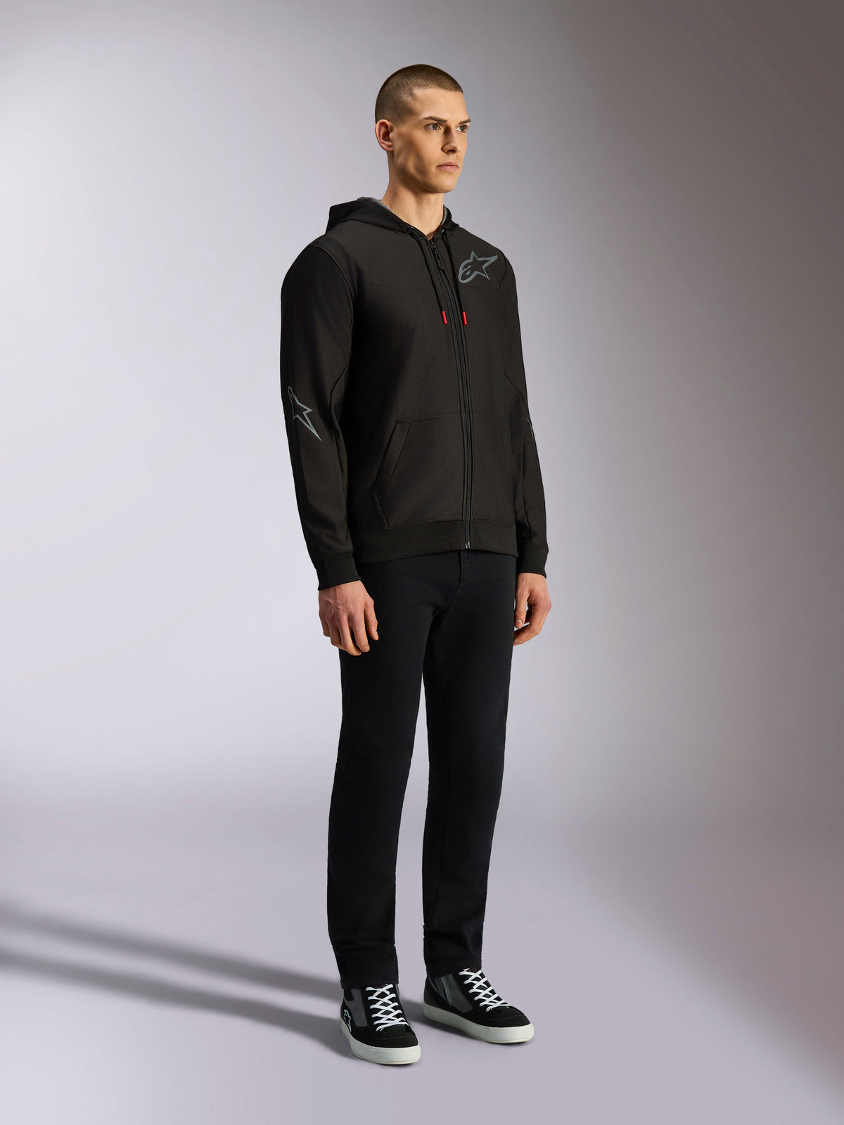 Felpa con cappuccio Alpinestars Sessions V3, streetwear casual da motorsport, felpa nera con zip e logo a stella grigio sul petto e sulla manica, cordini con punte rosse, polsini e orlo a coste, indossata da un modello con pantaloni neri e sneaker.