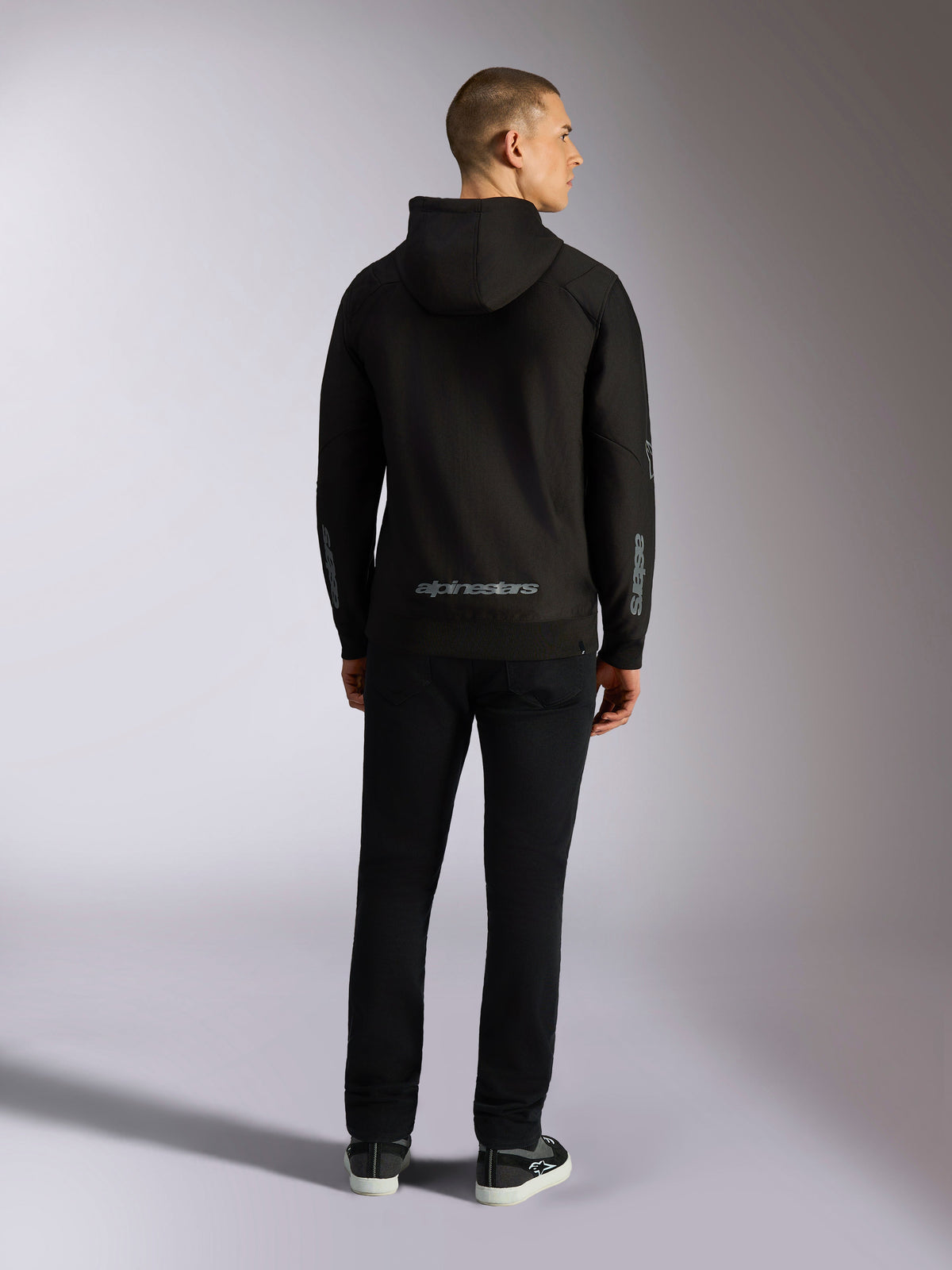 Felpa con cappuccio Alpinestars Sessions V3, streetwear casual, nera con scritta Alpinestars grigia sulla parte inferiore della schiena e marchio 'astars' sulle maniche, vista posteriore di un modello maschile che indossa la felpa e pantaloni neri, design moderno ispirato al motorsport