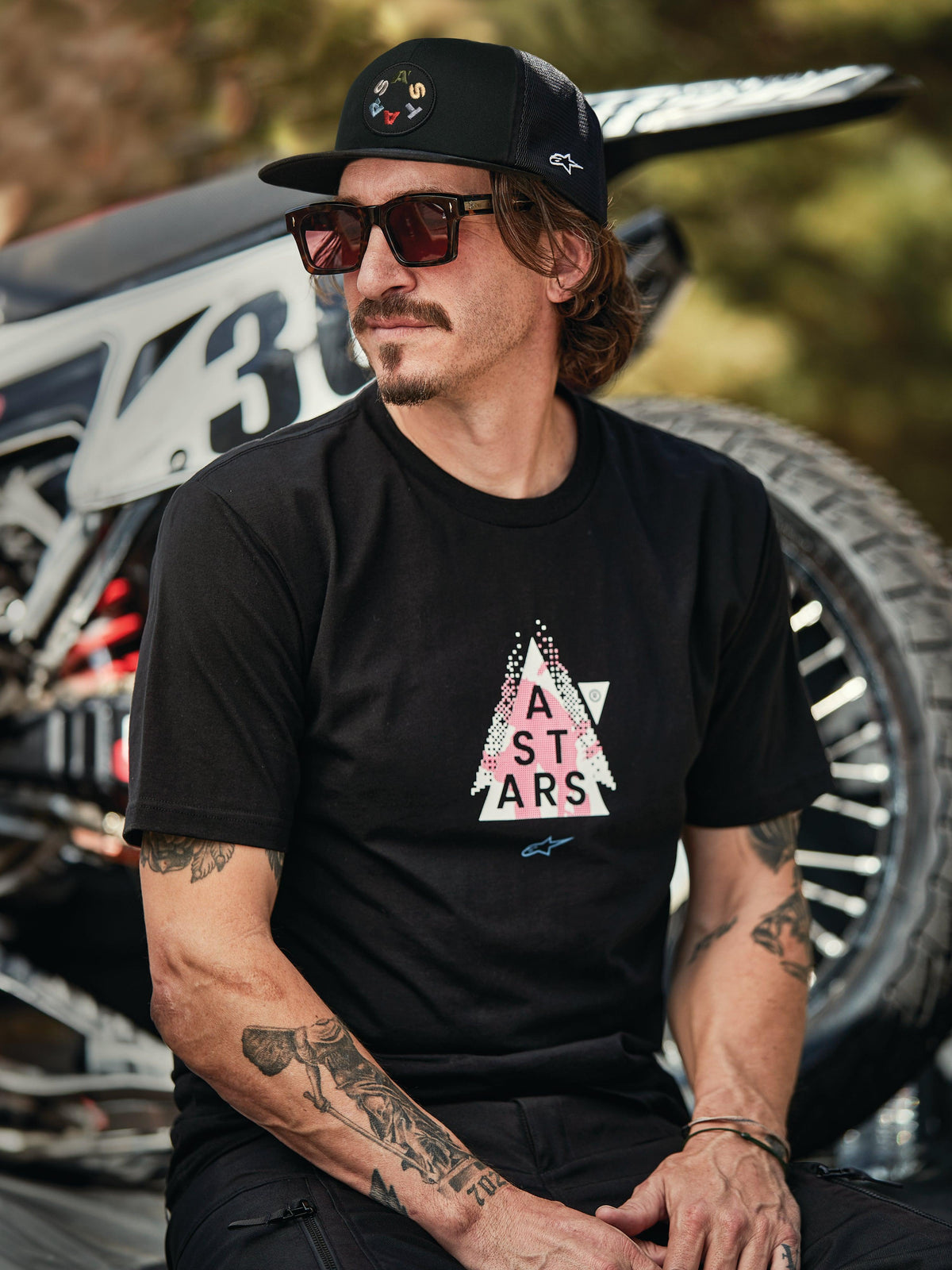 Cappello Alpinestars Gambit Trucker nero con patch logo circolare, indossato con T-shirt Alpinestars Soloist SS nera, scatto lifestyle con un uomo con occhiali da sole e tatuaggi seduto davanti a una moto da cross.