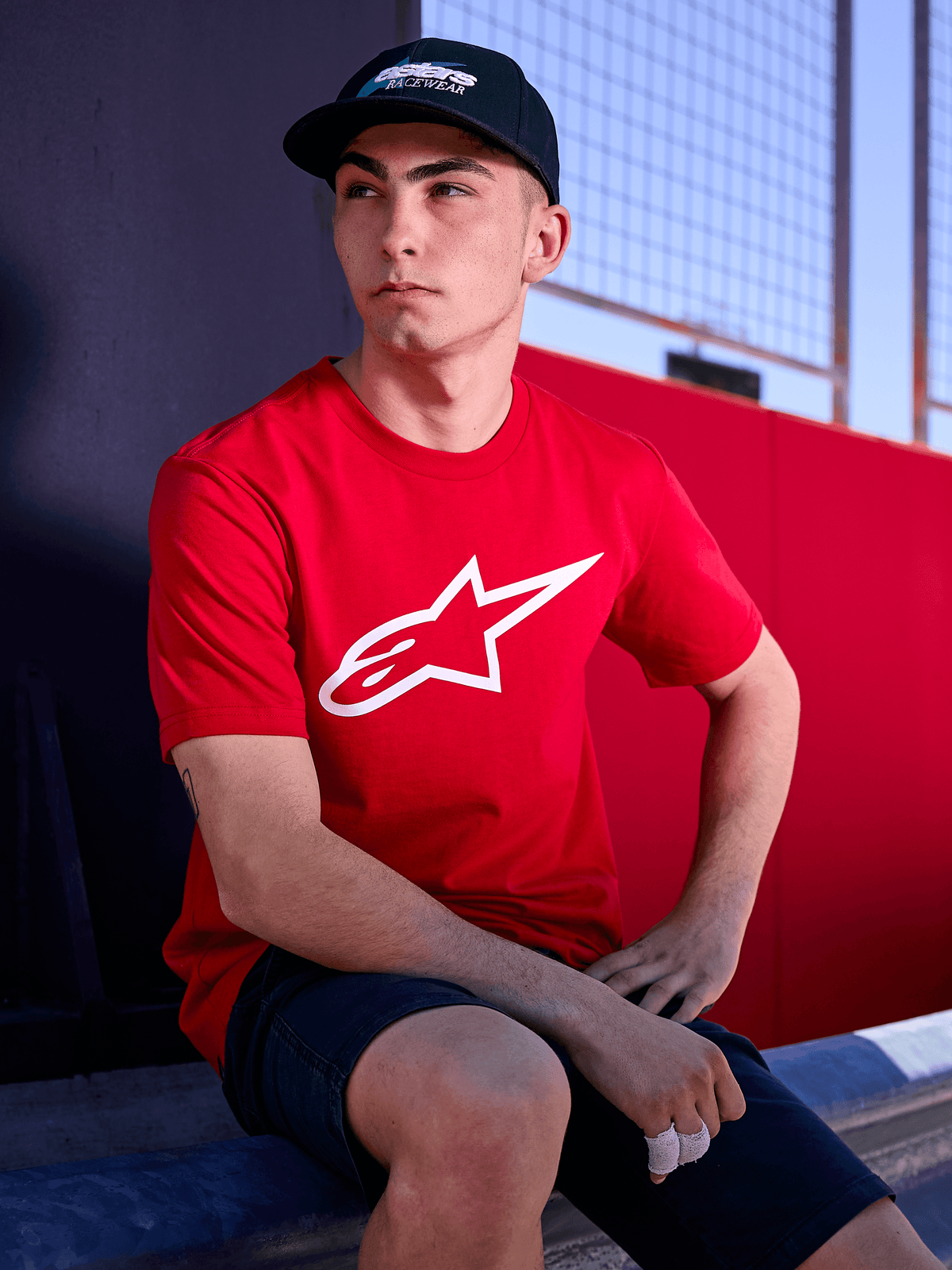 Cappello Profile Alpinestars nero, scatto lifestyle di un uomo che indossa il cappello con visiera curva e una t-shirt rossa con logo Alpinestars bianco, streetwear casual ispirato al motorsport